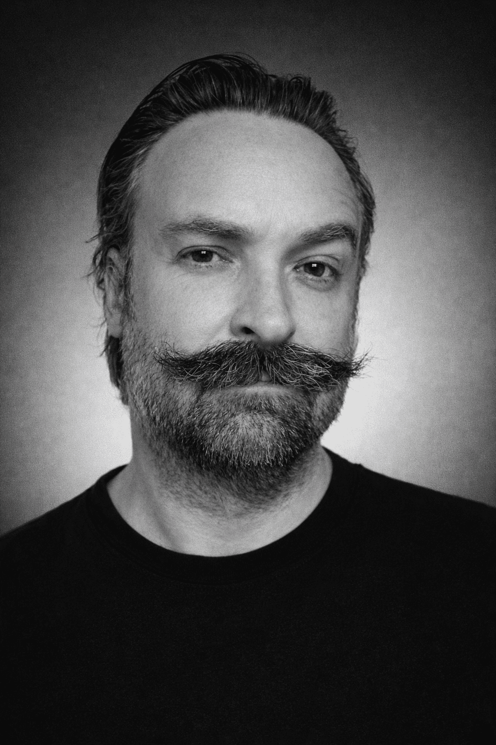 Man with glasses, beard, and tattoos wearing a black t-shirt against a green background.