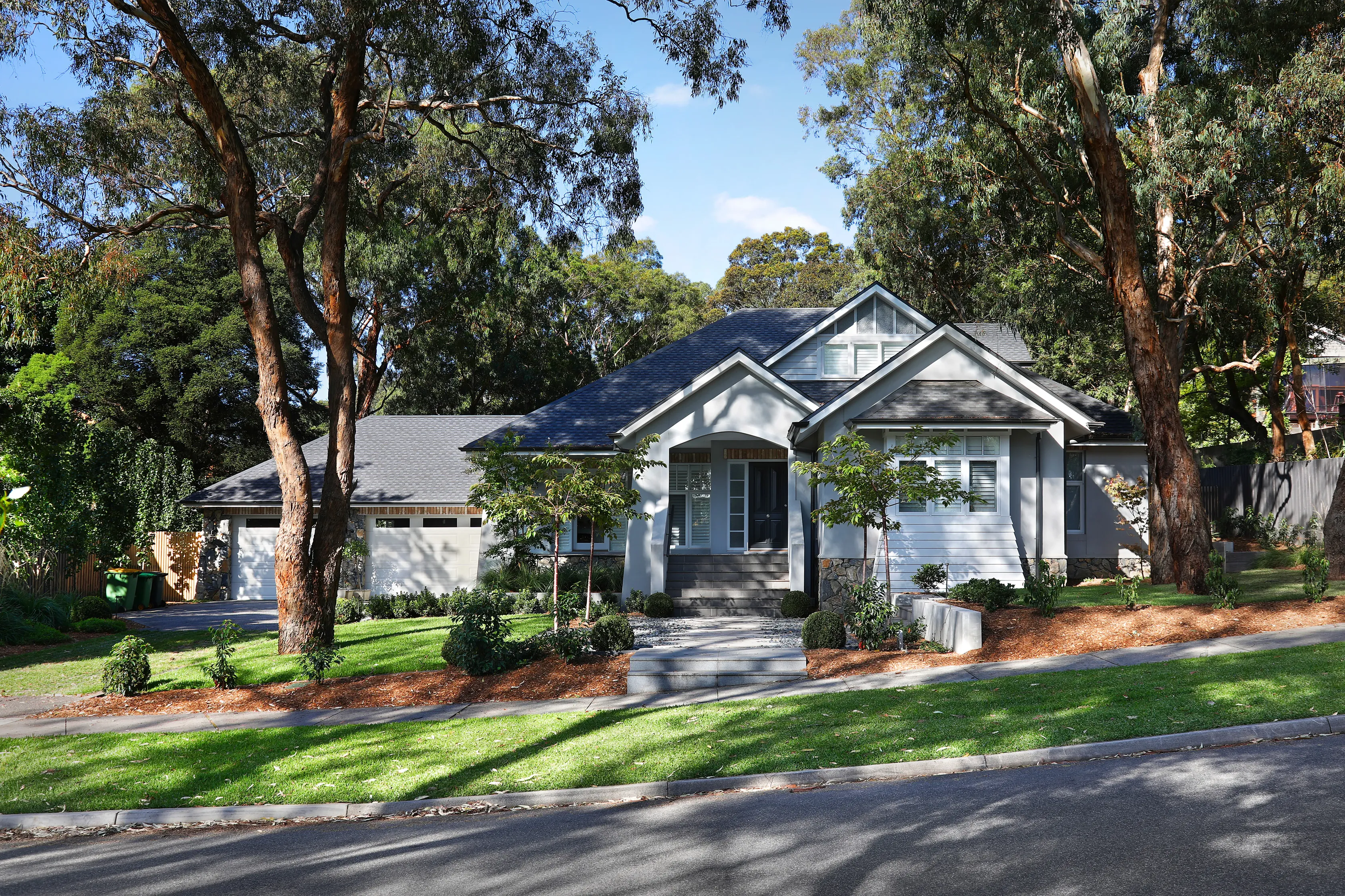 Street view of the Montmorency Residence, a single-storey rendered home with stacked-stone base, steep grey shingle roof and gables, set among tall gum trees on a landscaped lawn