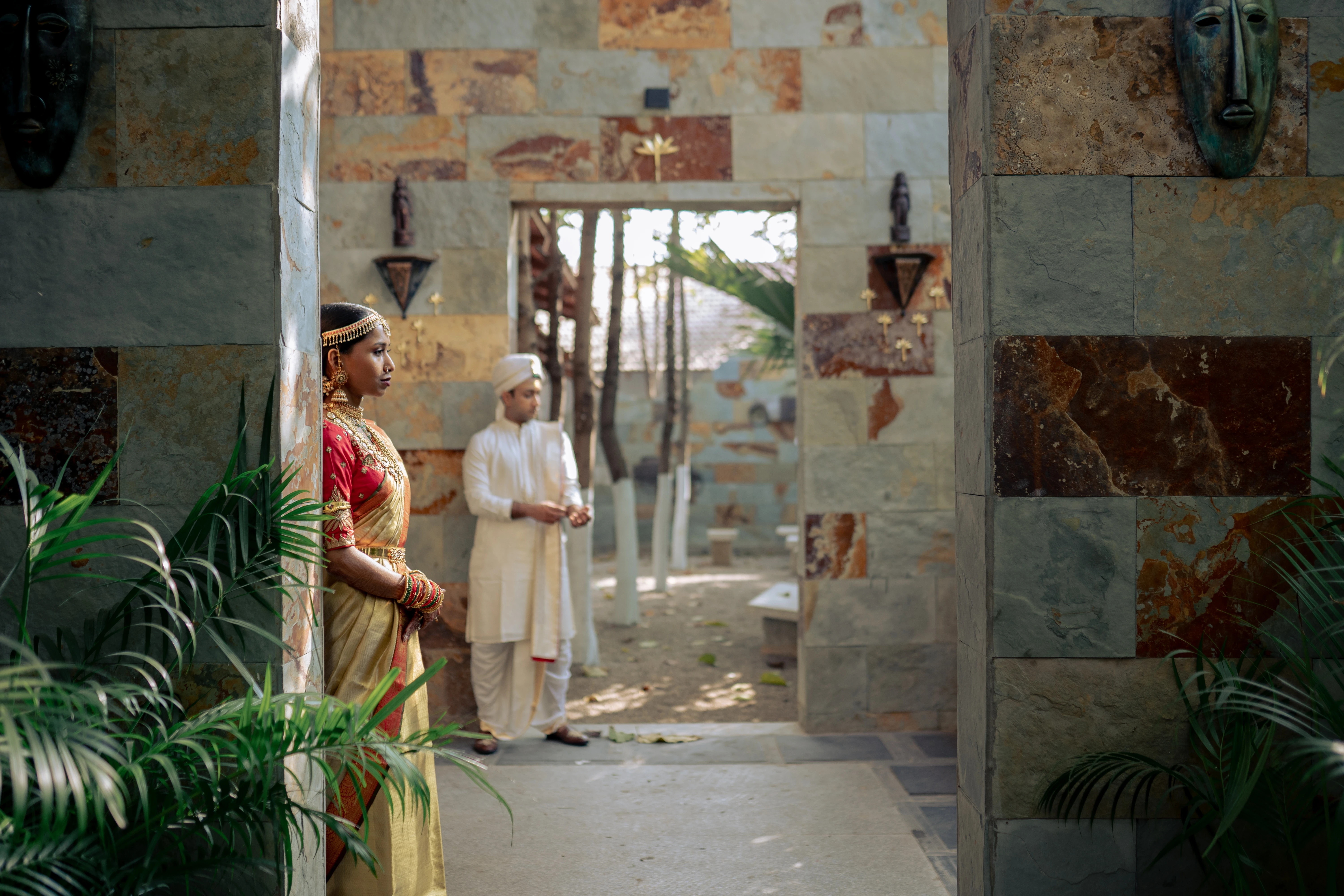 Vaishna in a red and gold sari leans against a stone wall, with Pranav in an ivory sherwani standing in the background, photographed by Out of The Blues, specialists in fine art wedding photography in Hyderabad.