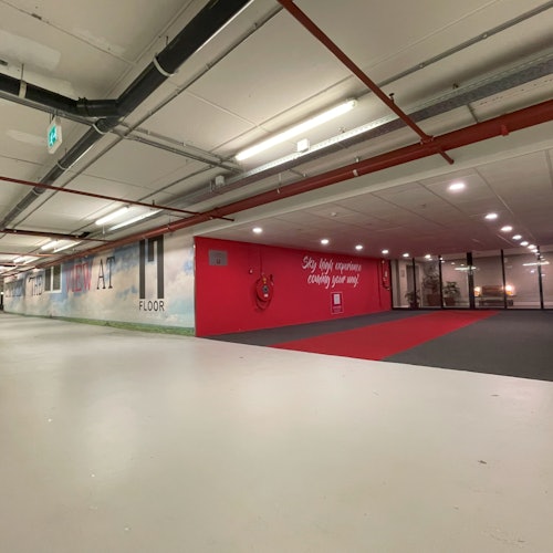 室内有一片宽敞的空旷区域，天花板上露出的管道和灯具，左墙上有一幅壁画，右侧铺着红色地毯。