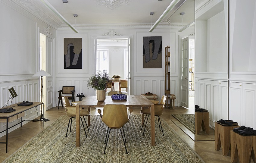 Bright Parisian room with ornate white wall paneling and parquet floor, furnished with a solid oak dining table surrounded by sculptural modern chairs on a textured rush-fiber rug; vases and design objects are arranged atop the table and adjacent console, creating a harmonious blend of architecture and artisan display.