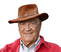 An elderly man wearing a brown leather cowboy hat and a red jacket smiles warmly at the camera. He stands in front of a wooden fence with greenery in the background, giving the image an outdoorsy, rustic feel. His friendly expression and casual attire convey a relaxed, approachable vibe.