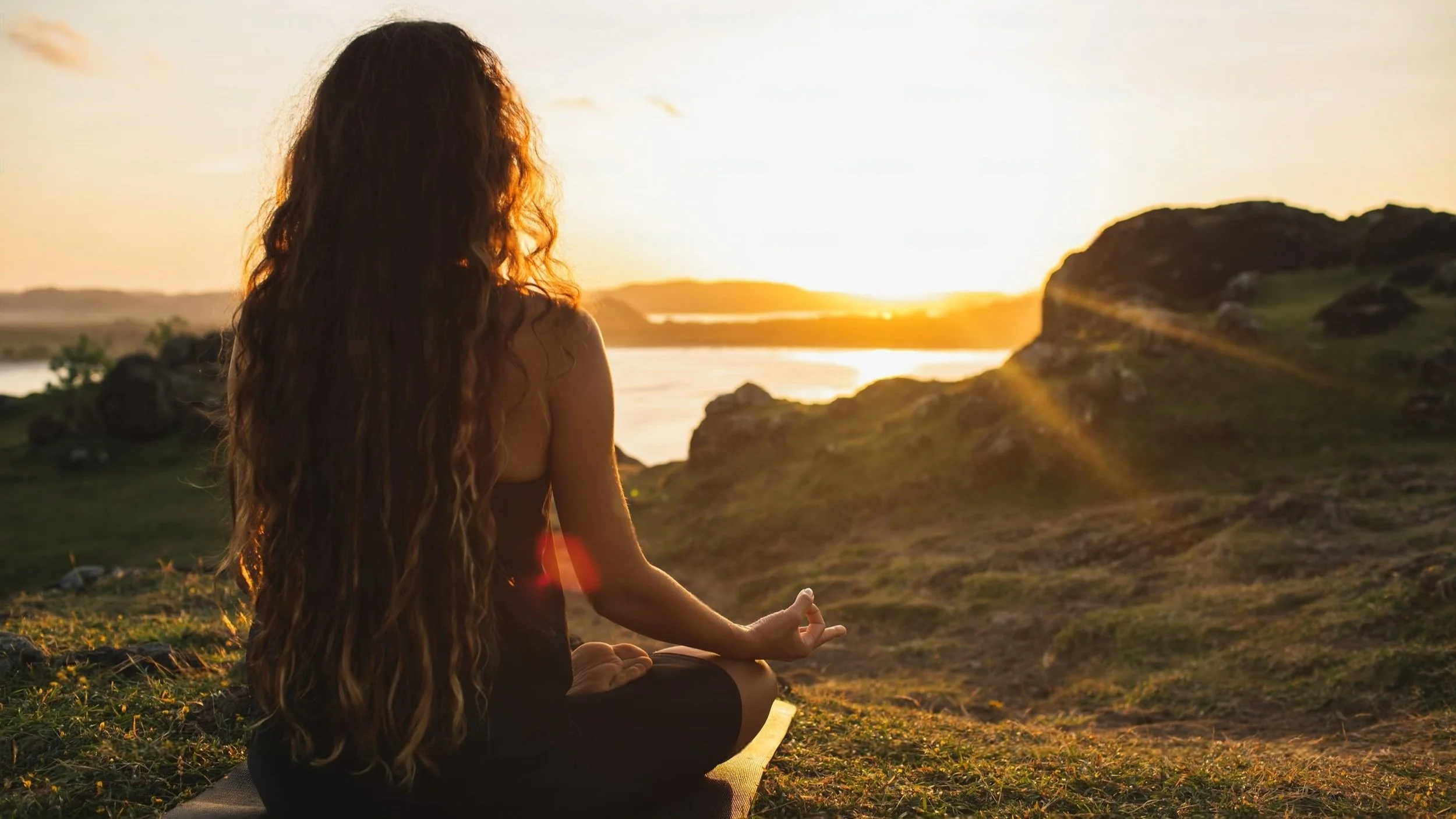 Person meditating outdoors at sunset, overlooking water and distant hills
