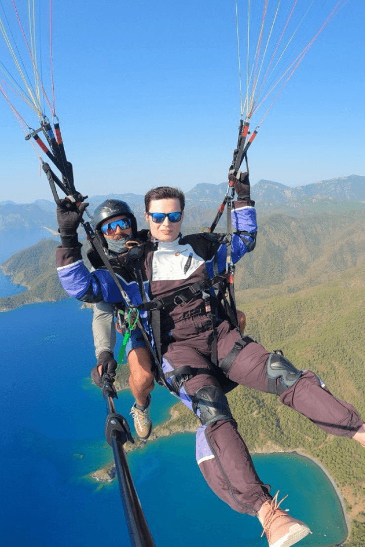 Two individuals are tandem paragliding over a stunning coastal landscape with vibrant blue waters, wearing protective gear and harnesses while enjoying the clear sky and scenic views beneath them.