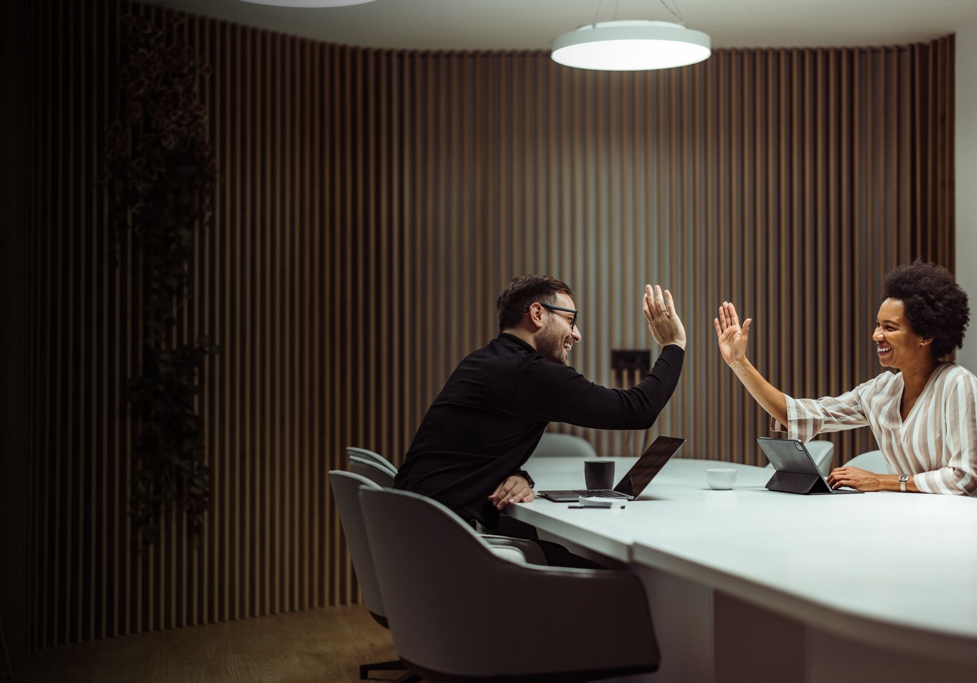 Man in glasses wearing a green shirt, writing in a notebook at a speckled table with a cup of coffee nearby, engaged in conversation.