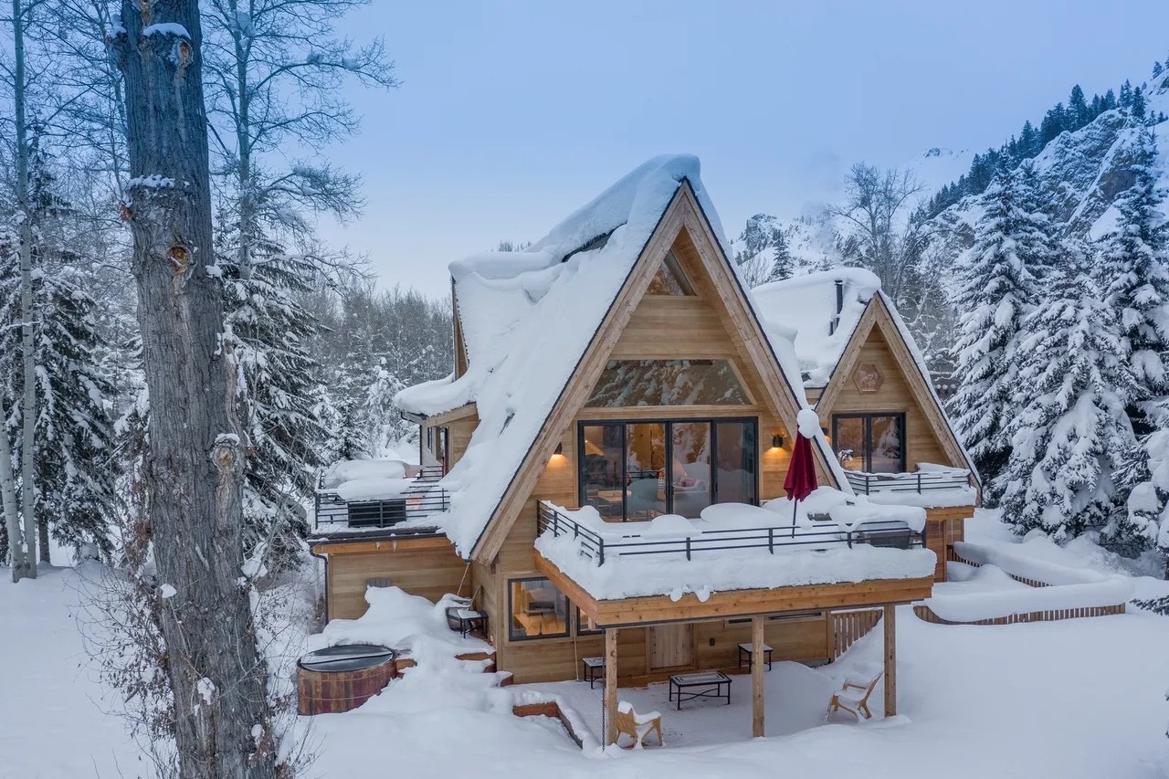 A-frame home in deep snow with a hot tub on the deck, surrounded by pine trees