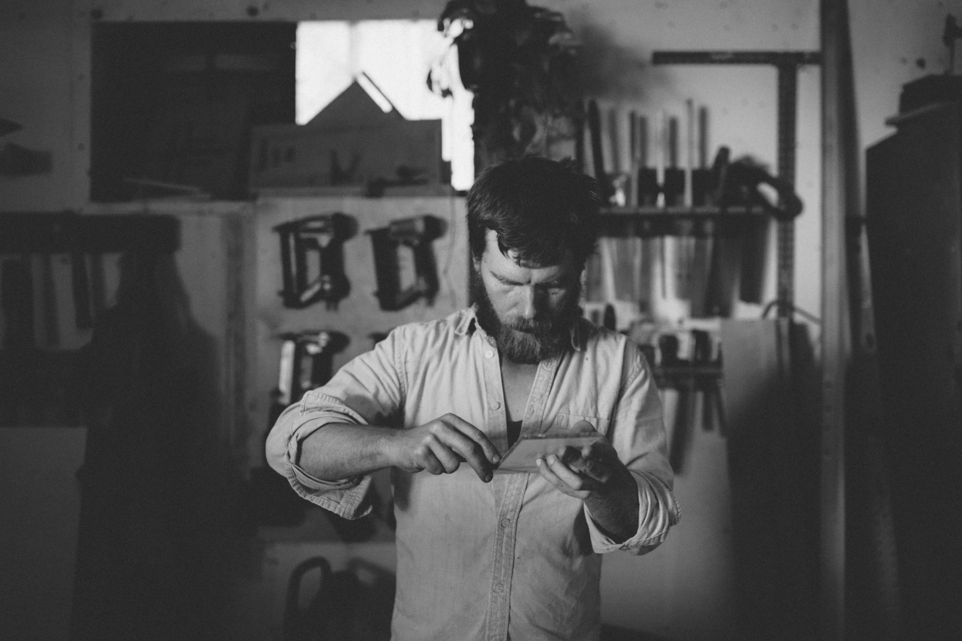 Ein Schreiner prüft konzentriert ein Holzwerkstück in seiner Werkstatt, umgeben von Werkzeugen und Materialien, aufgenommen in Schwarz-Weiß.