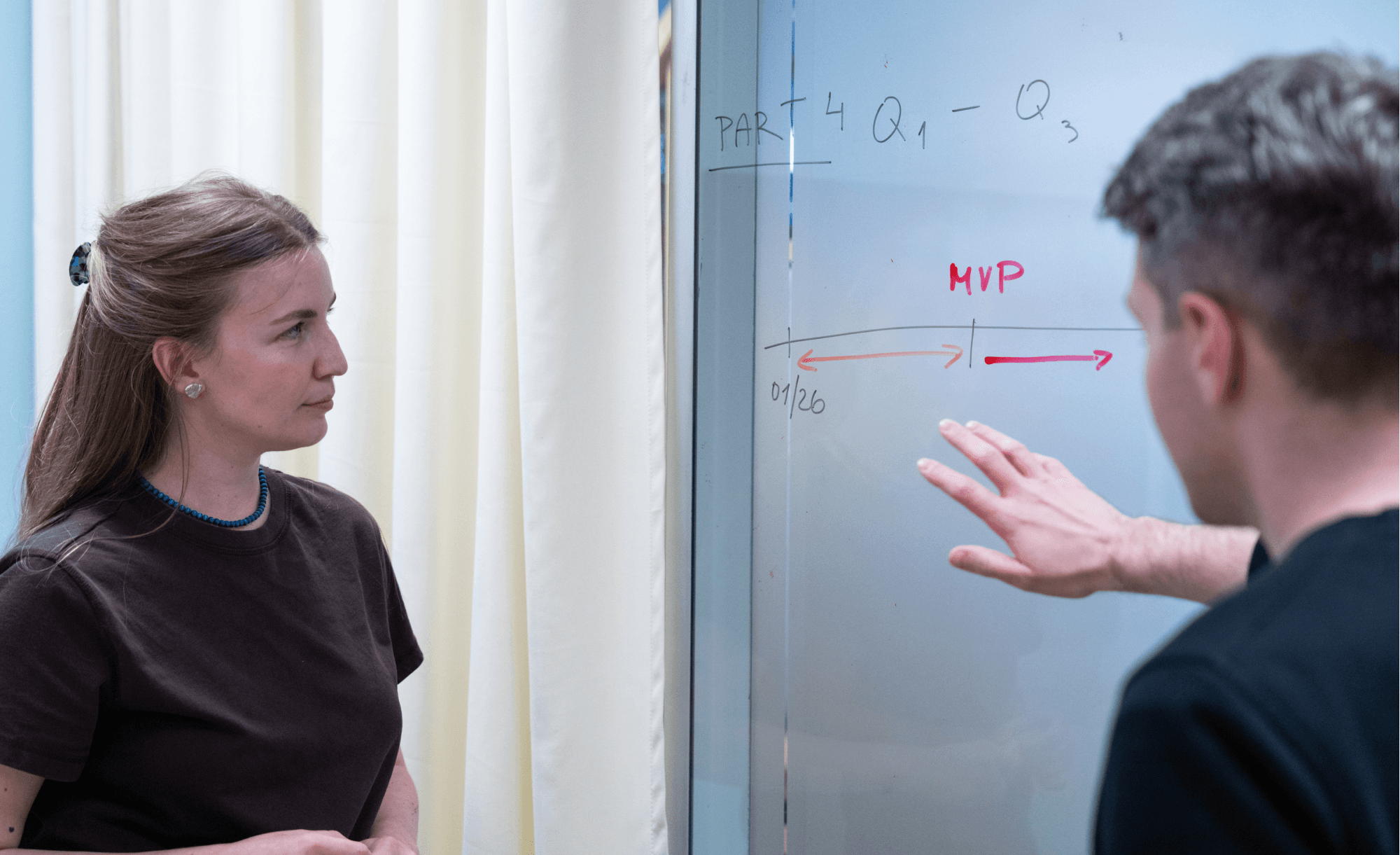 A product manager points at a whiteboard showing a release timeline marked "MVP" while a colleague listens.