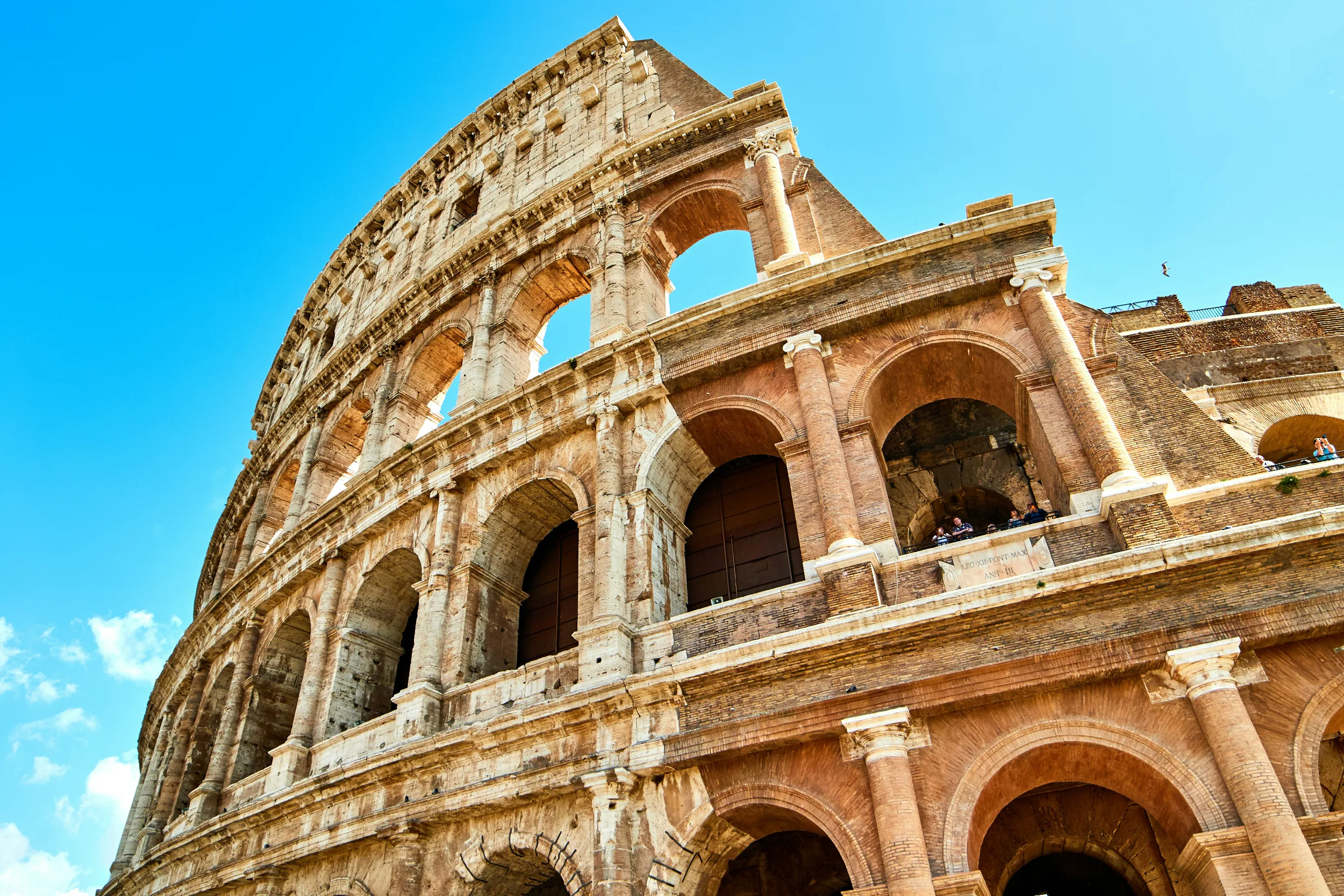 A closeup photo of the outside facade of the Roman Colosseum, highlighting its large arches. People are seen looking outside one of the archways. A blue sky shines down from above.