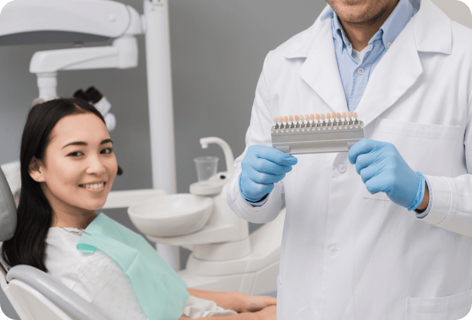 A smiling female patient sitting in a dental chair, while a dentist shows a shade guide for teeth whitening.