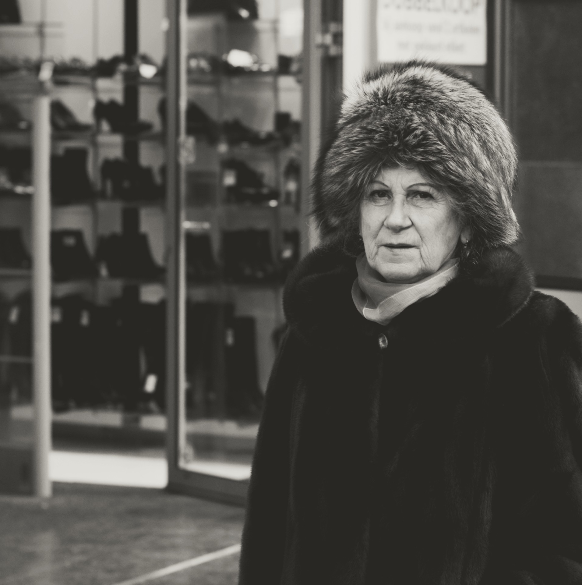 Black-and-white candid street photograph of an older individual in a fur coat and hat pausing before a shoe store display, highlighting textures, reflections, and a documentary urban mood.
