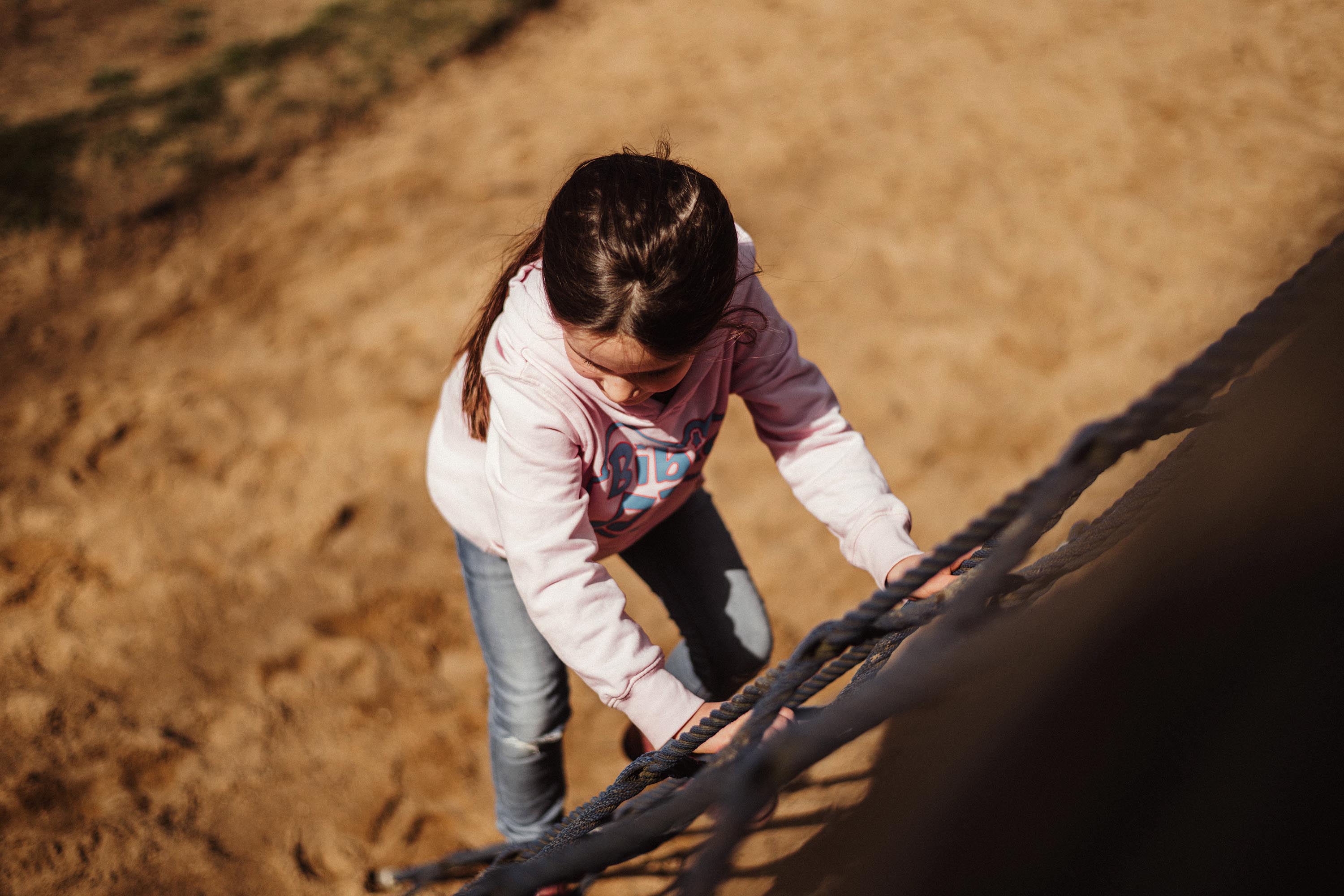 Authentische Kindergartenfotografie Gevelsberg: Ein junges Mädchen klettert auf einem Kletternetz hoch. Aufnahme von oben mit Bokeh-Hintergrund von PK photography 4 Kids NRW.