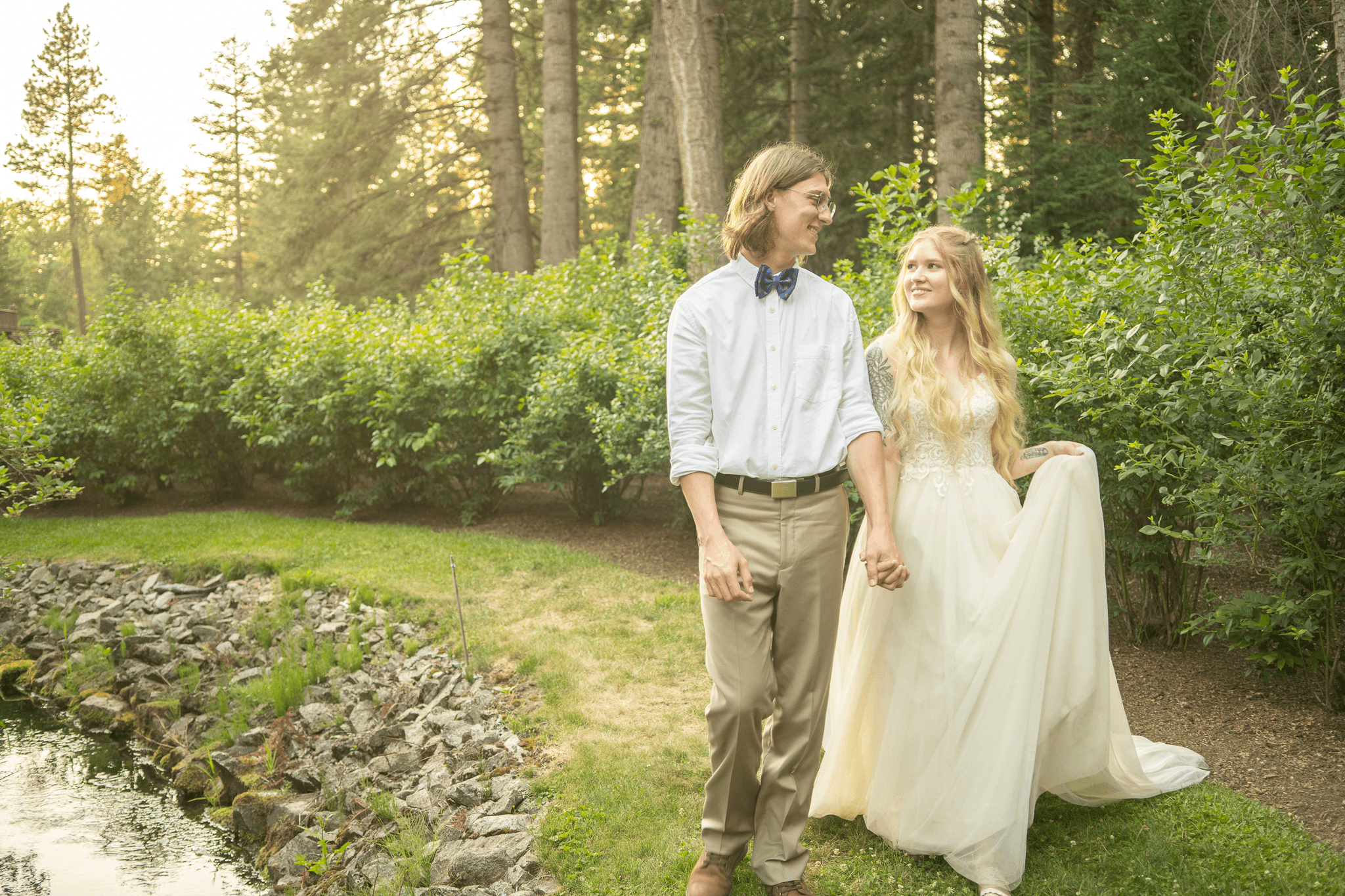 A beautiful couple walking through a garden with a stream to their left and beautiful foliage and trees behind them