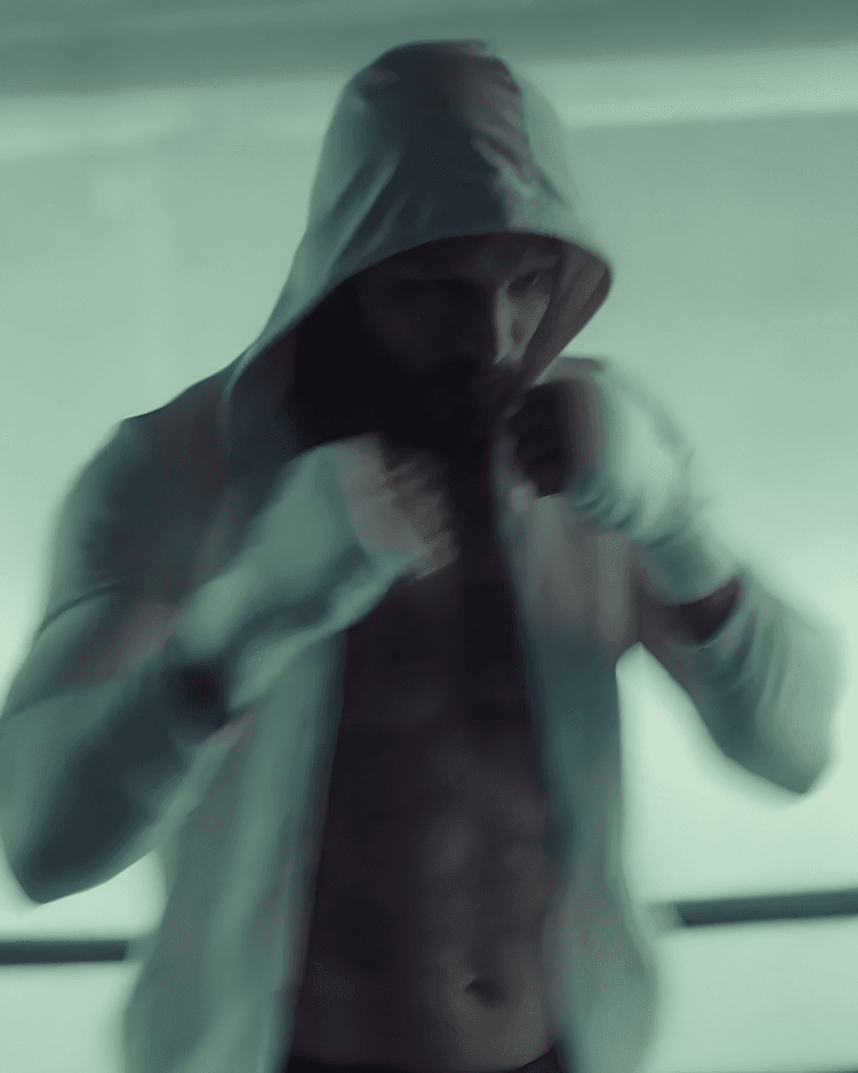 A blurred image of a hooded individual in motion, wearing hand wraps, suggesting a boxing or martial arts setting.