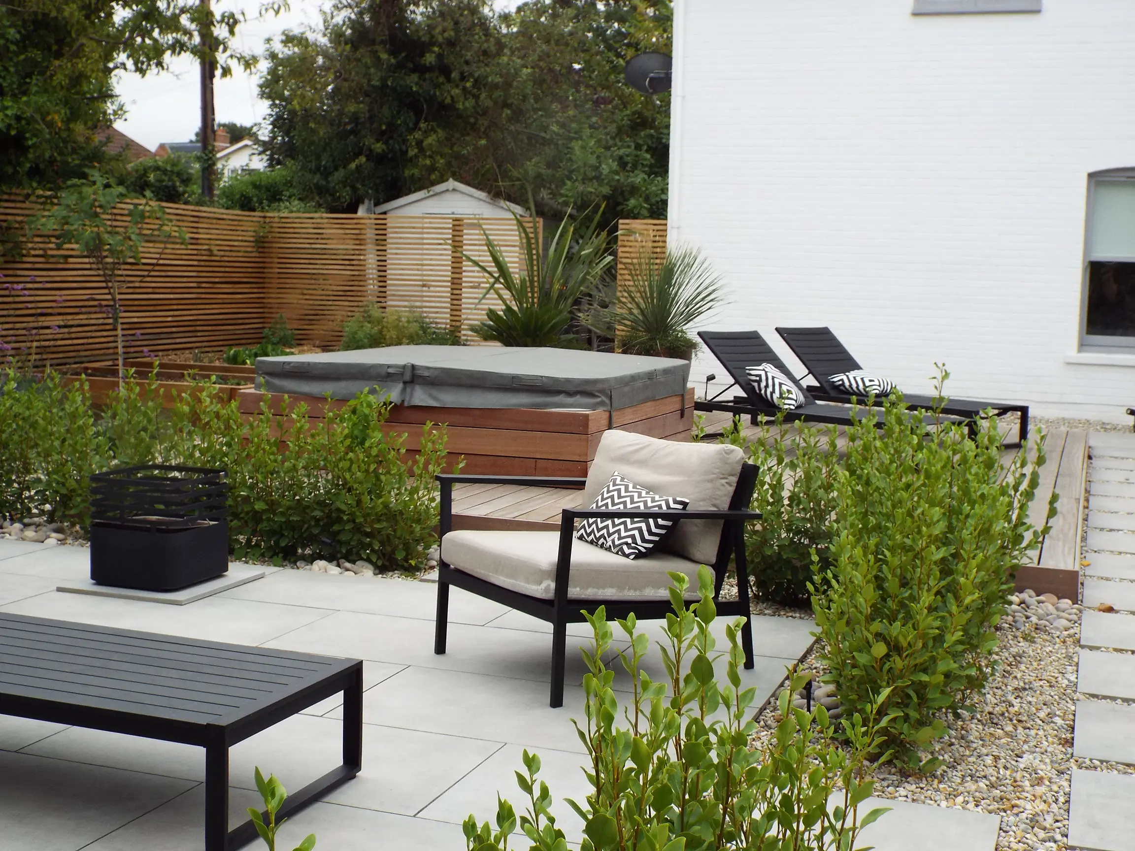 A modern outdoor patio with lounge chairs, a hot tub, and greenery surrounding a sleek, minimalist design.