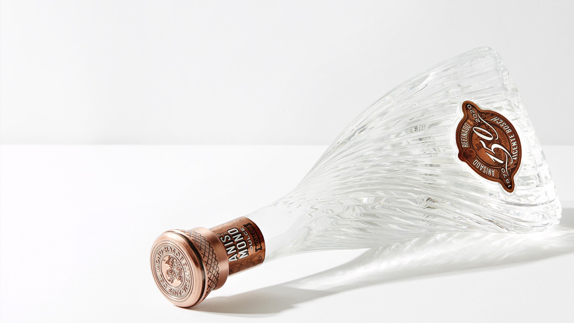 Elegant glass bottle with textured swirls lies on its side. Copper-toned cap and label read "ANIS NOVO". Minimalist and sophisticated ambiance.
