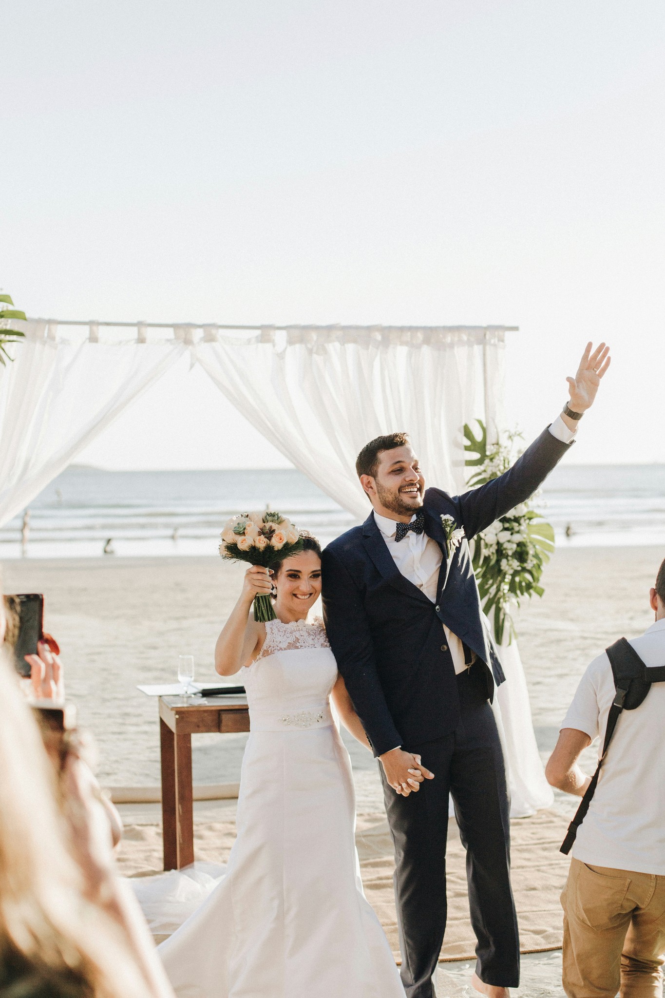 Newlyweds celebrating with raised hands and bouquet after their beach wedding ceremony with the ocean behind them