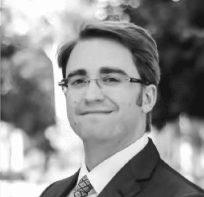 black and white photo of the upper part of a smiling man in a suit and white shirt without tie. This is the Chief Data Scientist of BeyondTrucks, Dr. John Carisson