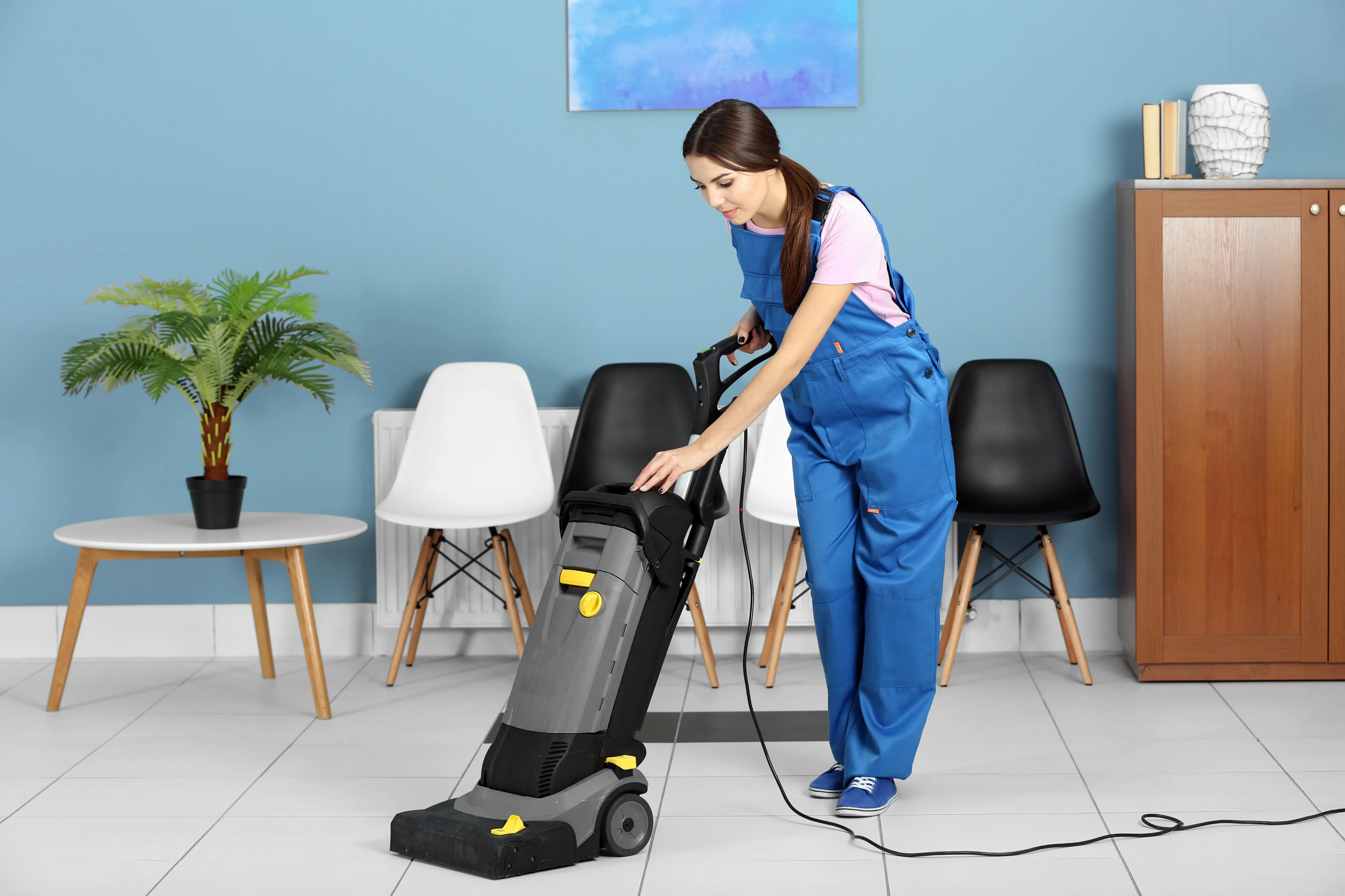 A professional female cleaner in a blue uniform using a heavy-duty industrial vacuum cleaner on a white floor in a modern, brightly lit living room.