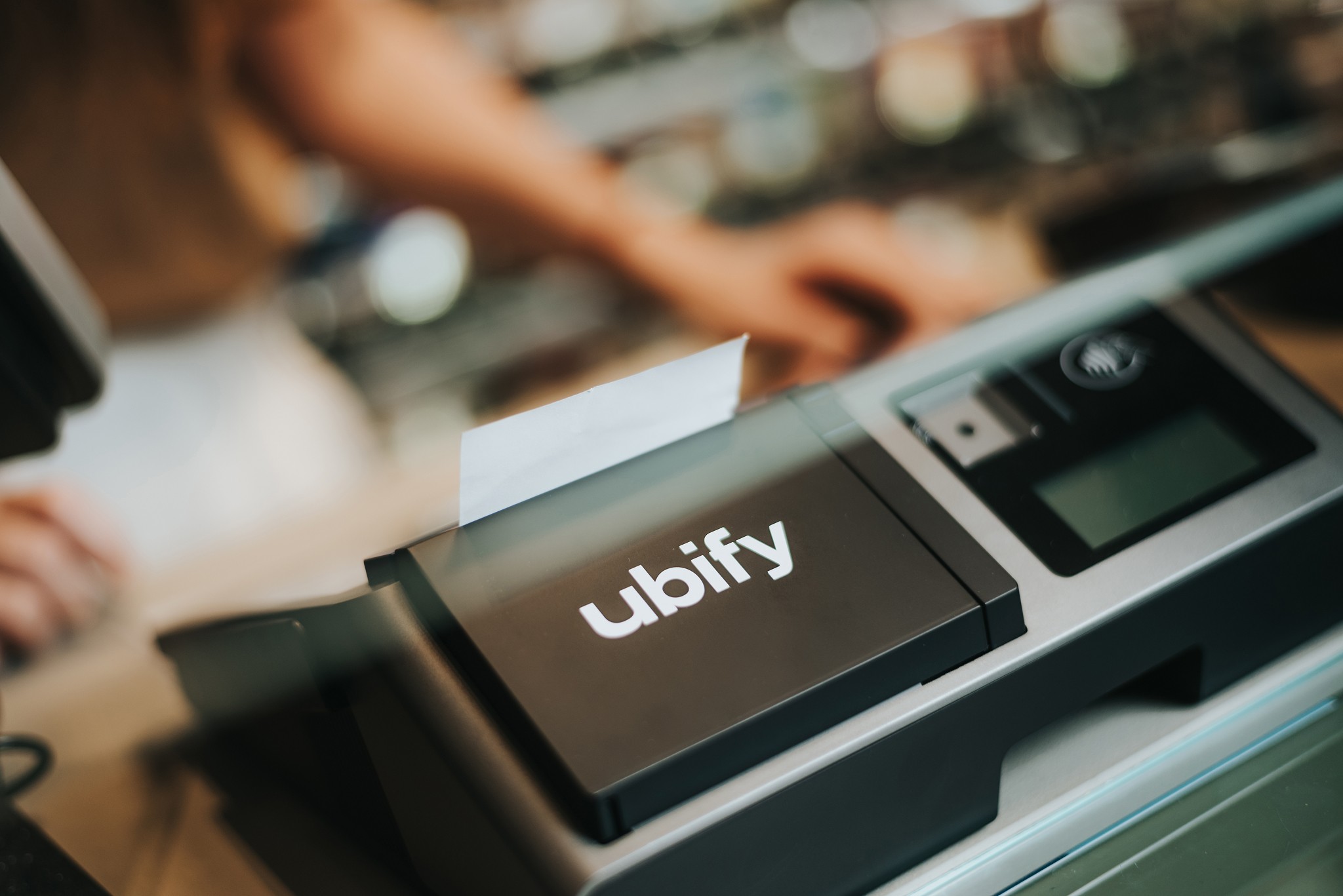 Cassa Ubify in primo piano con scontrino stampato, posizionata su bancone di negozio con negoziante sullo sfondo.
