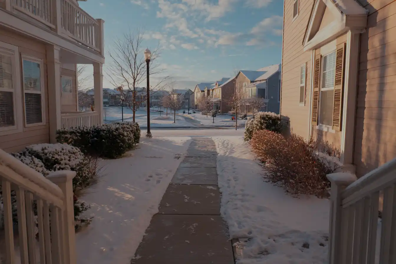 Snow-covered suburban street at sunrise. Sidewalk leads between two houses surrounded by snowy shrubs, under a clear blue sky with fluffy clouds.