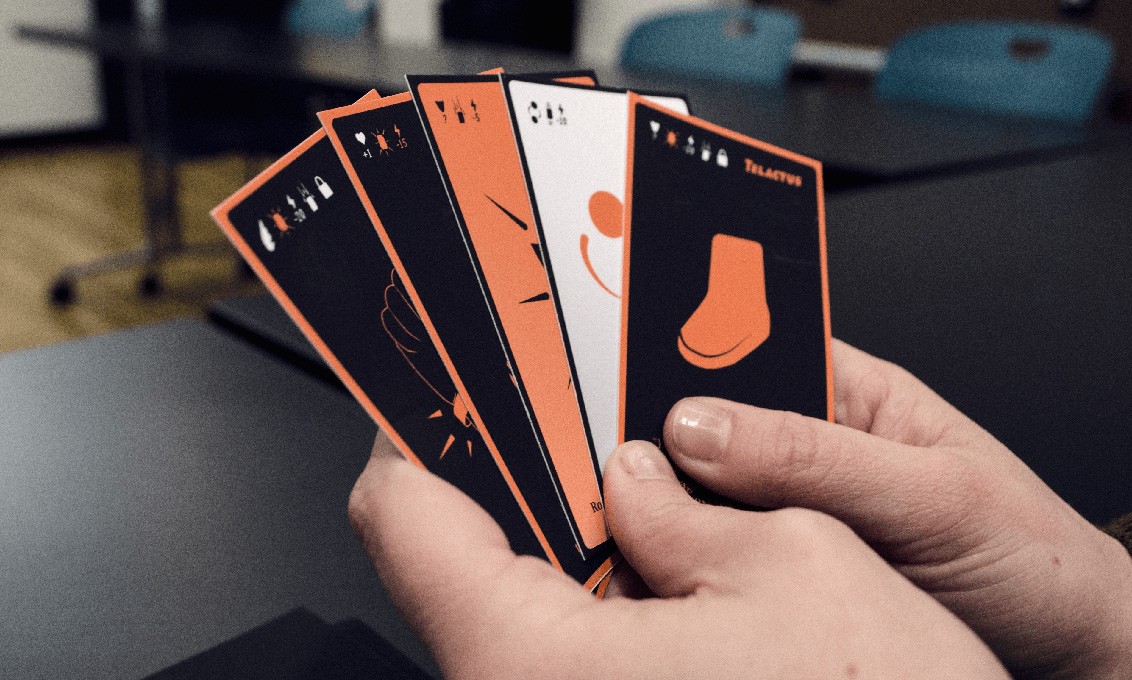 A hand holds four cards with black and orange designs. The top card displays a white background with an orange boot silhouette. The remaining three cards feature black designs with orange outlines and text, including “Talantuo” at the top right. The cards are held over a dark table, with a blurred background showing office chairs and tables.