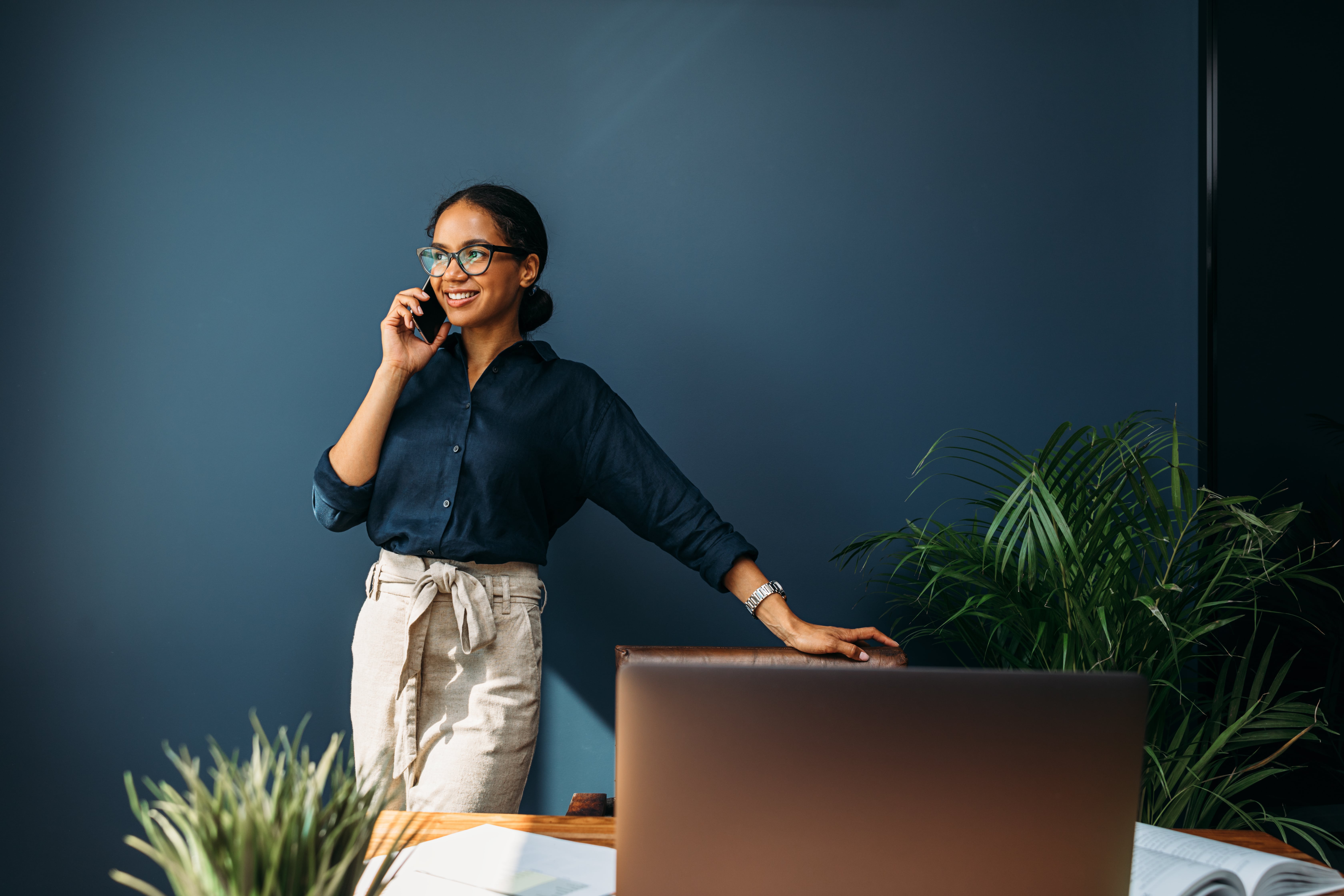 Een vrouw in een donker shirt staat bij een bureau en houdt een telefoon aan haar oor, met planten op de achtergrond.
