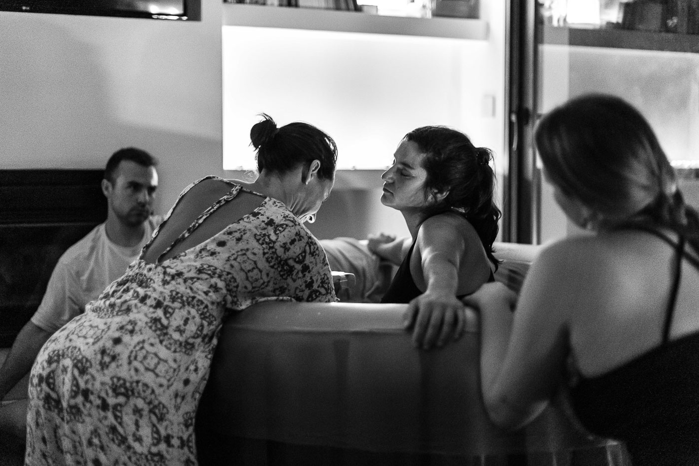 Mujer en trabajo de parto en casa, acompañada por su pareja y doula, parto en piscina inflable, fotografía documental en blanco y negro