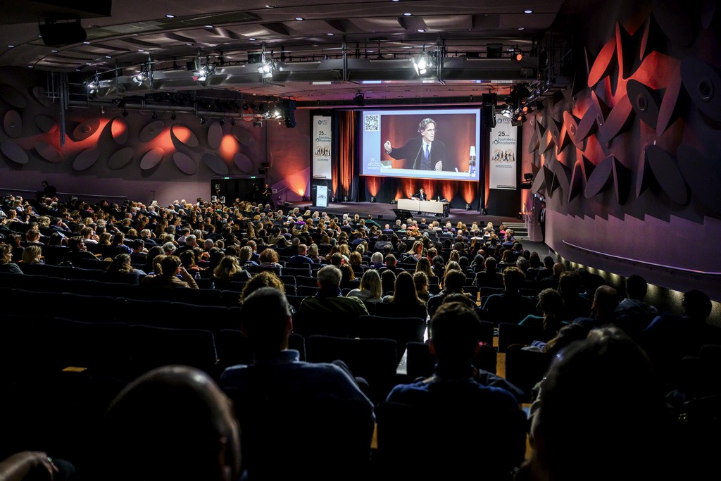 Salle de conférence pleine de spectateurs dans la pénombre avec le conférencier éclairé et sur écran géant.