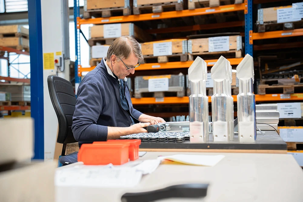 Ein Mitarbeiter von RITTER Starkstromtechnik montiert Bauteile an einem Arbeitstisch in der Fertigung. Im Hintergrund sind Regale mit Material und Komponenten für die Produktion zu sehen.