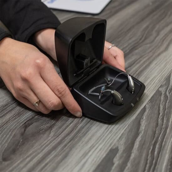 A hand opens a black box revealing a pair of wireless earbuds on a wooden surface.