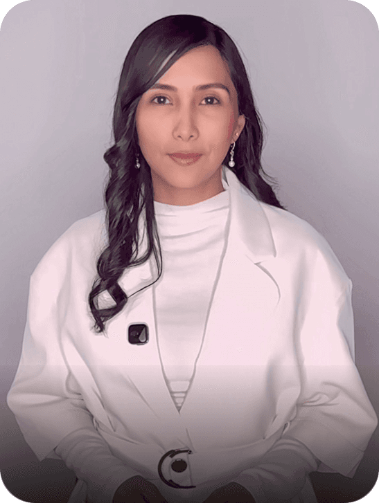 A smiling person with curly hair sits at a desk, looking at a laptop against a dark background.