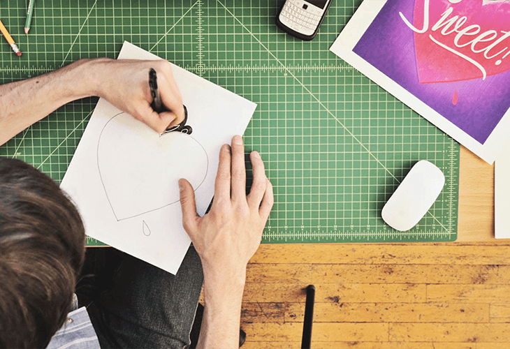 A person is seated at a green cutting mat, drawing on a white sheet of paper with a black marker. The paper contains a heart shape outlined in black ink, with a small drop of ink visible near the bottom left corner. The person’s left hand holds the marker, while their right hand rests on the paper. A smartphone, a white eraser, and a framed print with a pink and purple heart design are visible on the mat. The mat has grid lines marking inches. The person is wearing dark-colored pants, and the mat rests on a light brown wooden surface.