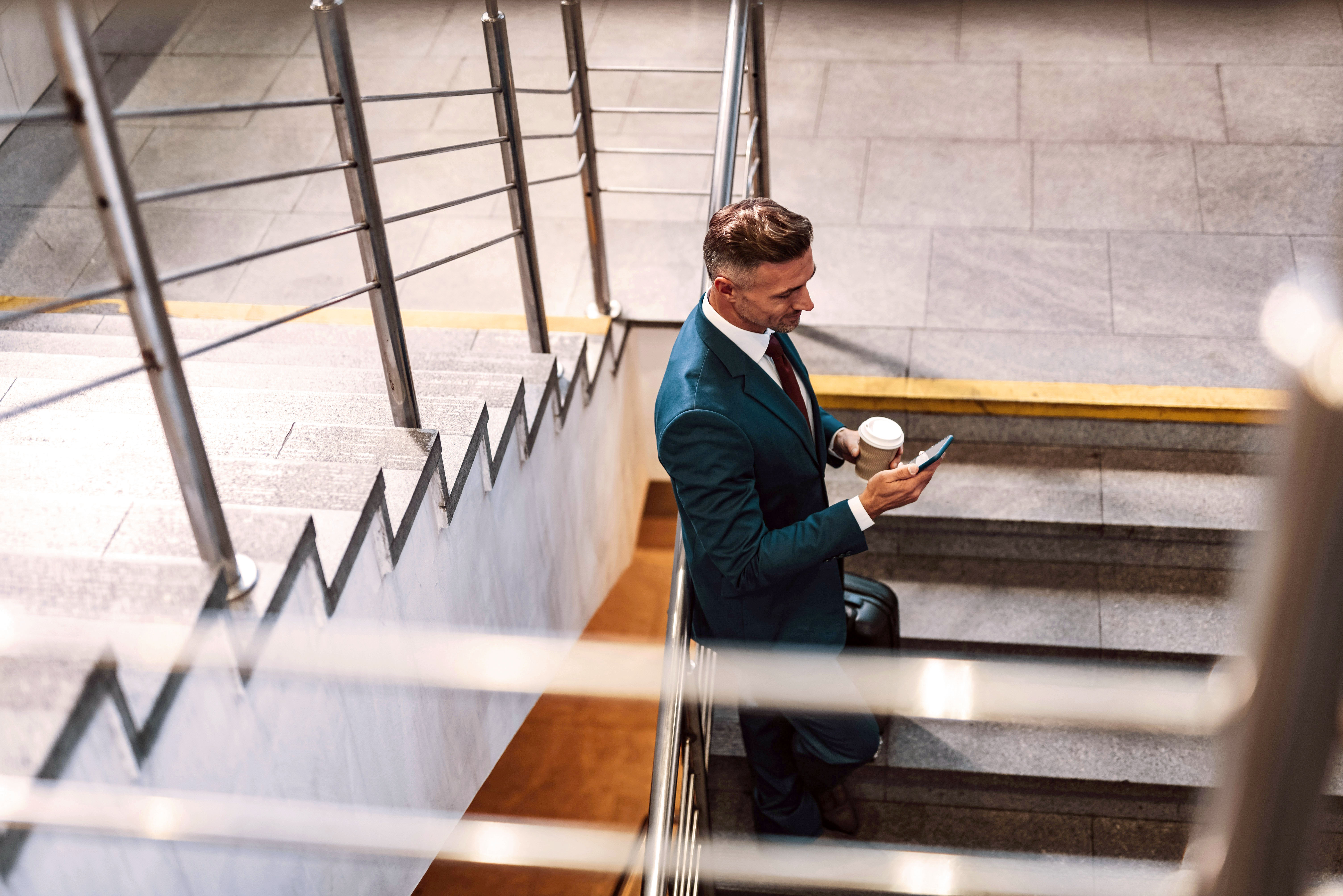 A person in a suit holds a cup while standing on stairs, looking up in a modern indoor setting.