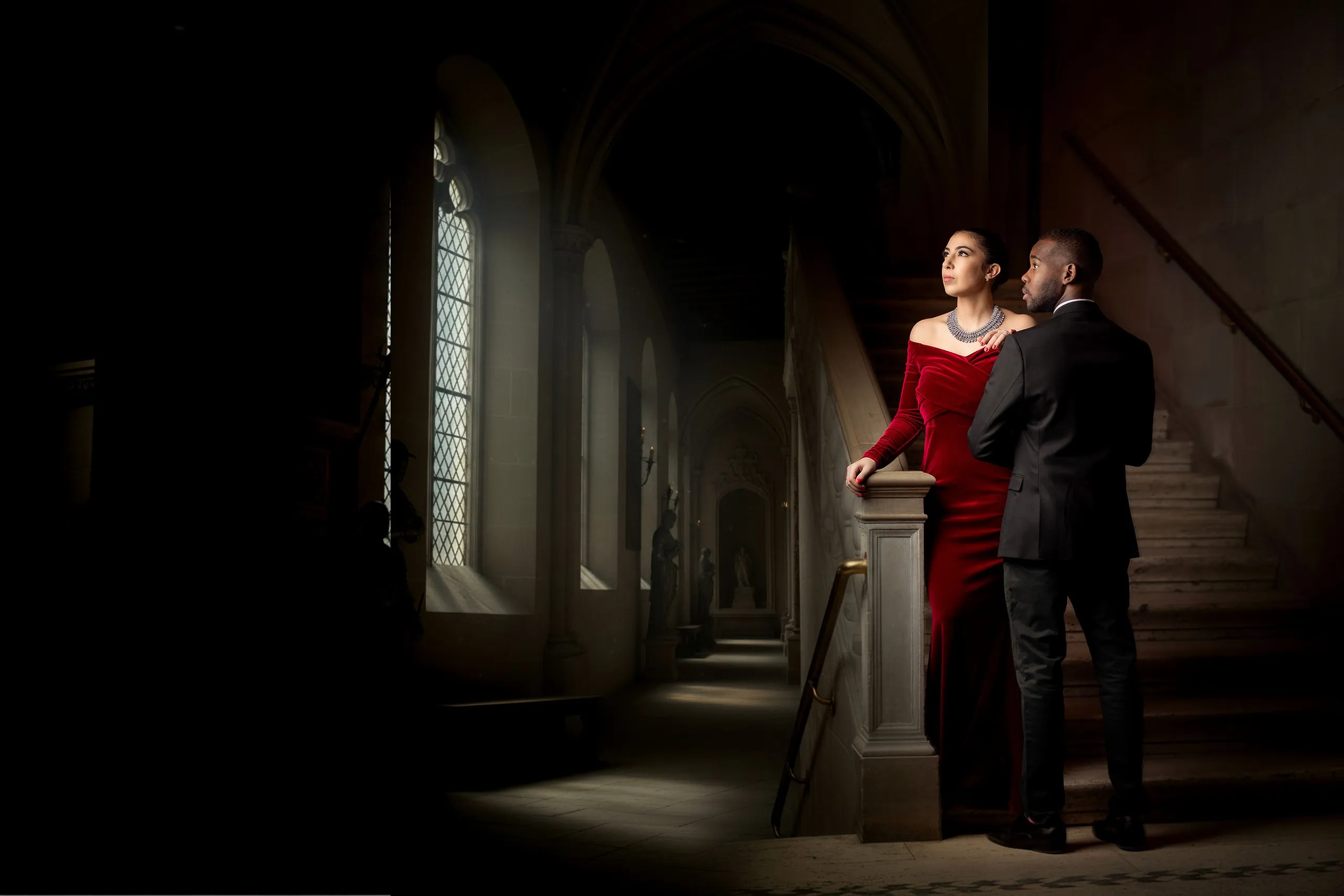 Deux personnes élégamment vêtues posant dans un couloir orné, avec une femme en robe rouge et un homme en costume noir, se tenant près d'une balustrade.