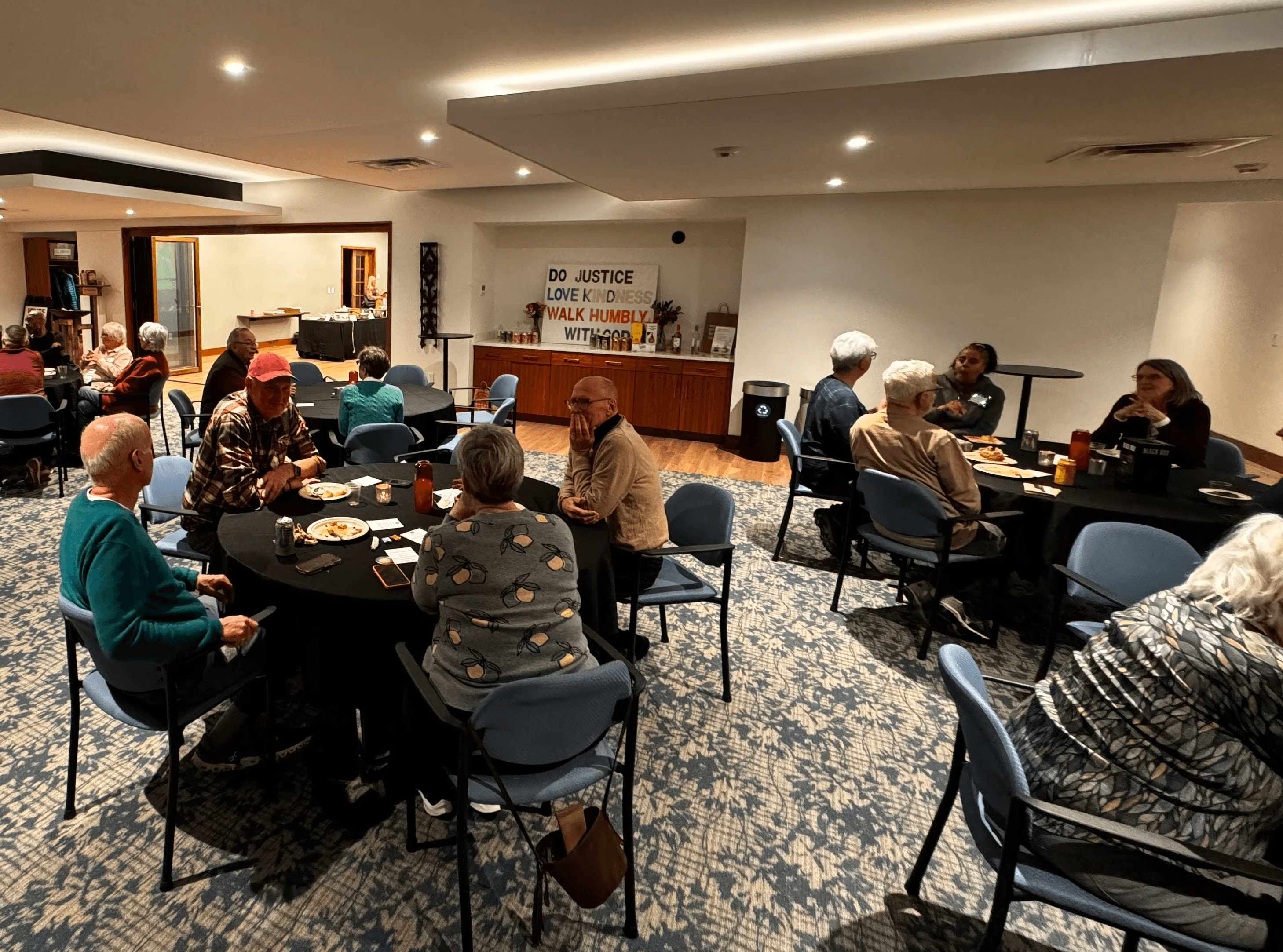 A group of people seated at tables enjoying conversation and food in a community room at Grace Trinity Church, with a sign in the background reading ‘Do justice, love kindness, walk humbly with God’.