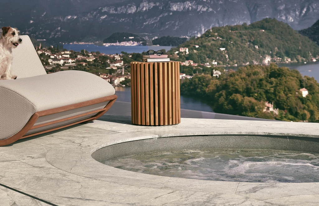 Round Layer side table in solid Iroko wood, composed of interlocked vertical panels in a radial pattern, styled in a luxury poolside setting with lounge chairs and tropical greenery – modern outdoor furniture with sculptural design.