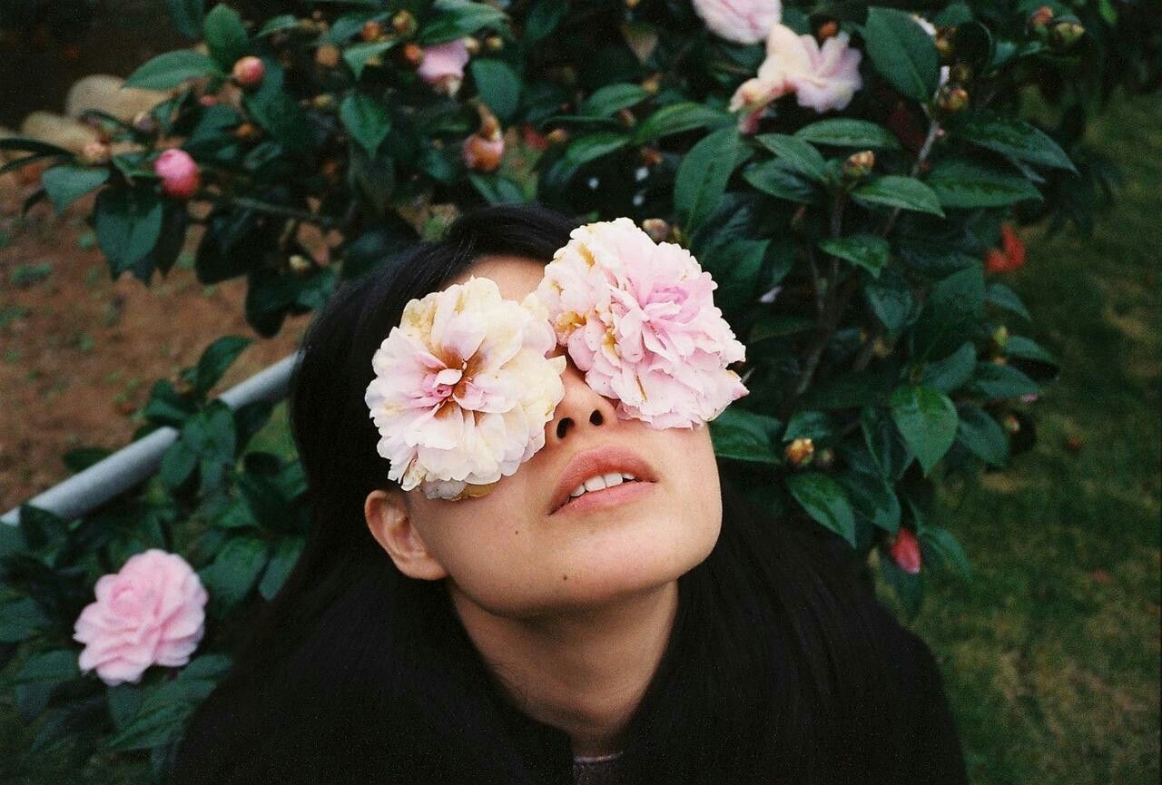 A black-haired woman gazing upward, with a flower placed over each eye.