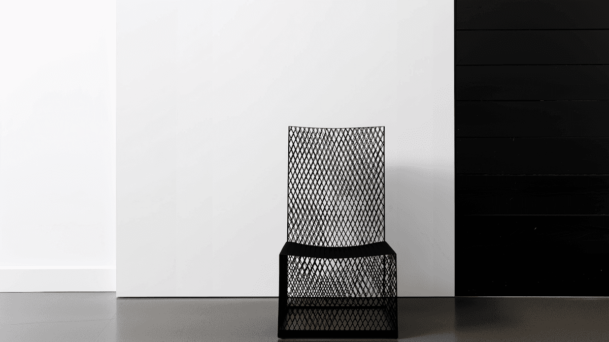 Minimalist black mesh chair with a geometric pattern, placed against a clean white wall and dark panel backdrop.