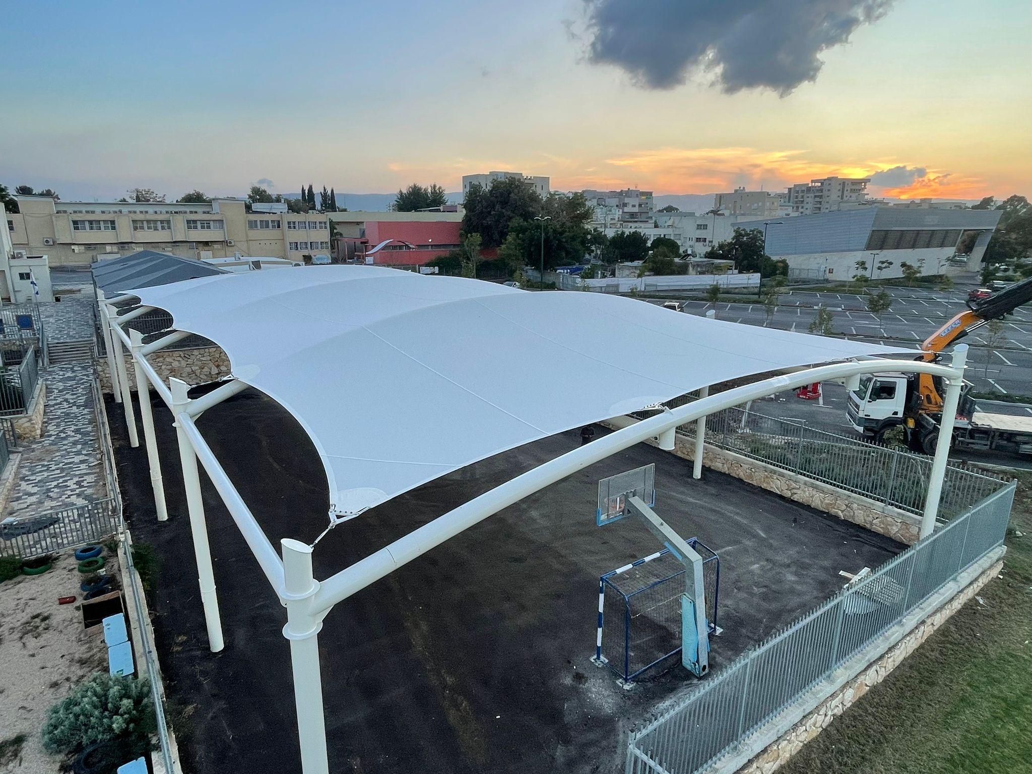 Large beige curved sunshade over an outdoor area with basketball hoop and fence in an urban setting at sunset.
