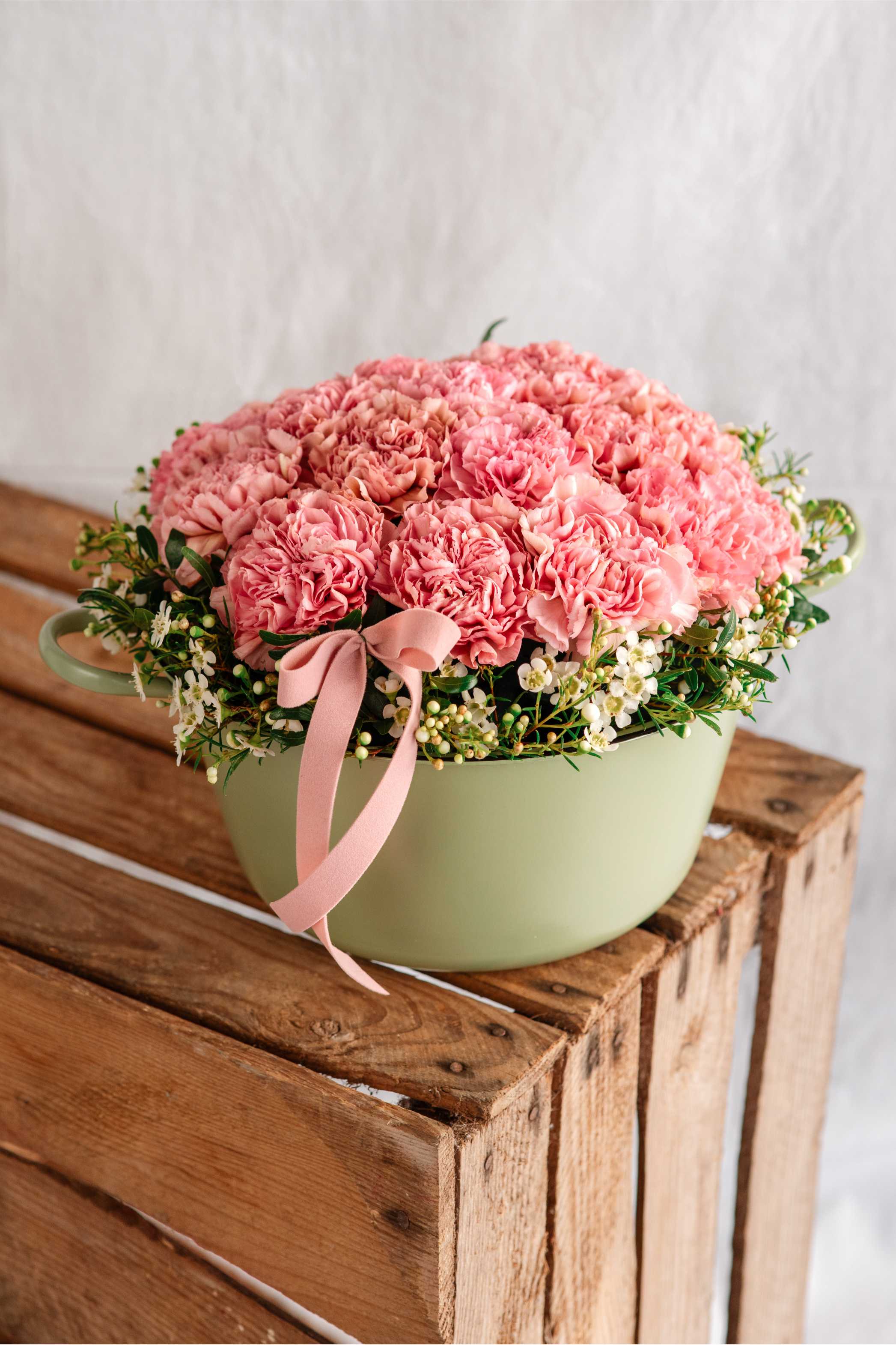 Composició de clavells rosats en una base de metall verd amb acabat mat, envoltada de flor blanca petita i cinta rosa, col·locada sobre una caixa de fusta natural.