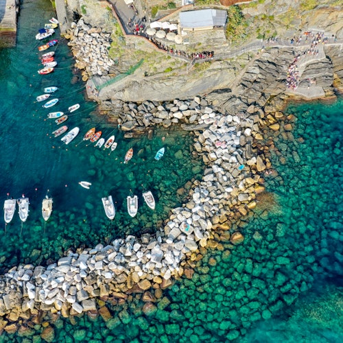 Letecký pohled na skalnaté pobřeží s malým přístavem plným lodí. Průzračná tyrkysová voda obklopuje skály a lodě.