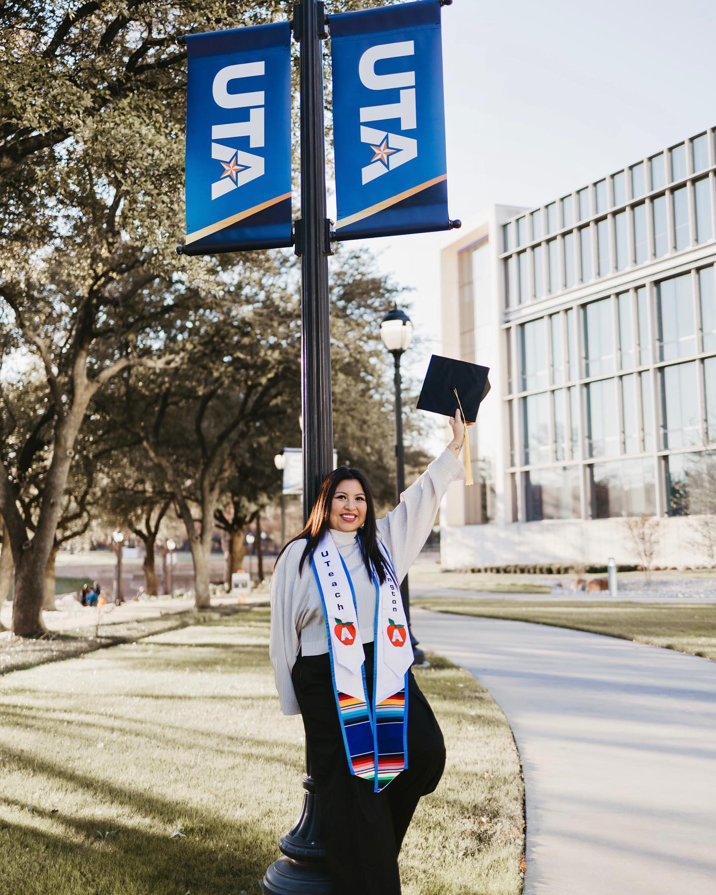 A proud graduate in a cap and gown stands on a university campus, with UTA banners hanging from a light post, joyfully raising her graduation cap. Her stole adorned with colorful cultural symbols suggests a celebration of her heritage and academic achievements.