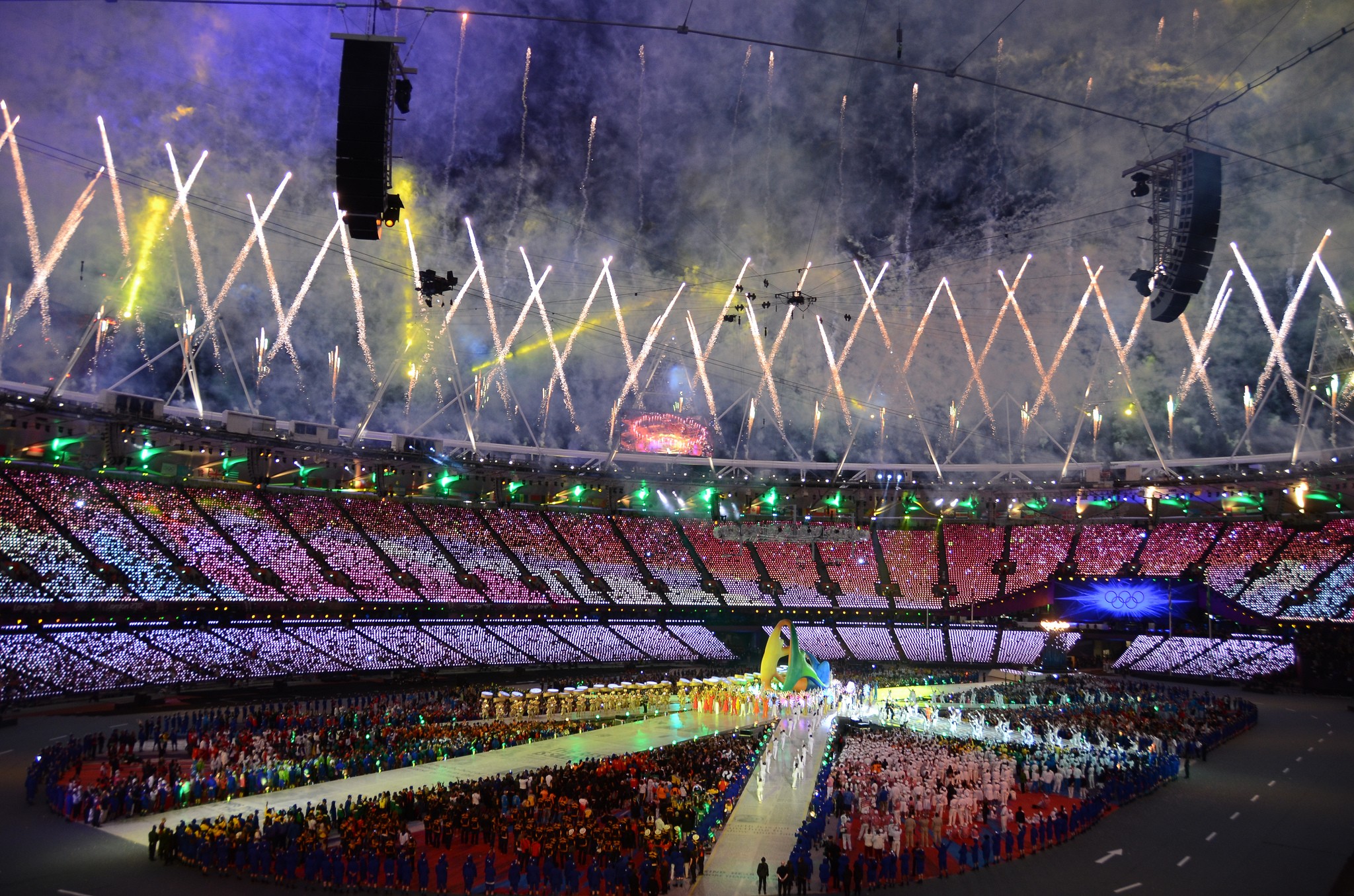 A vibrant stadium is illuminated with colorful lights and fireworks during a spectacular evening ceremony, with crowds gathered on the field, creating an atmosphere of celebration and excitement.
