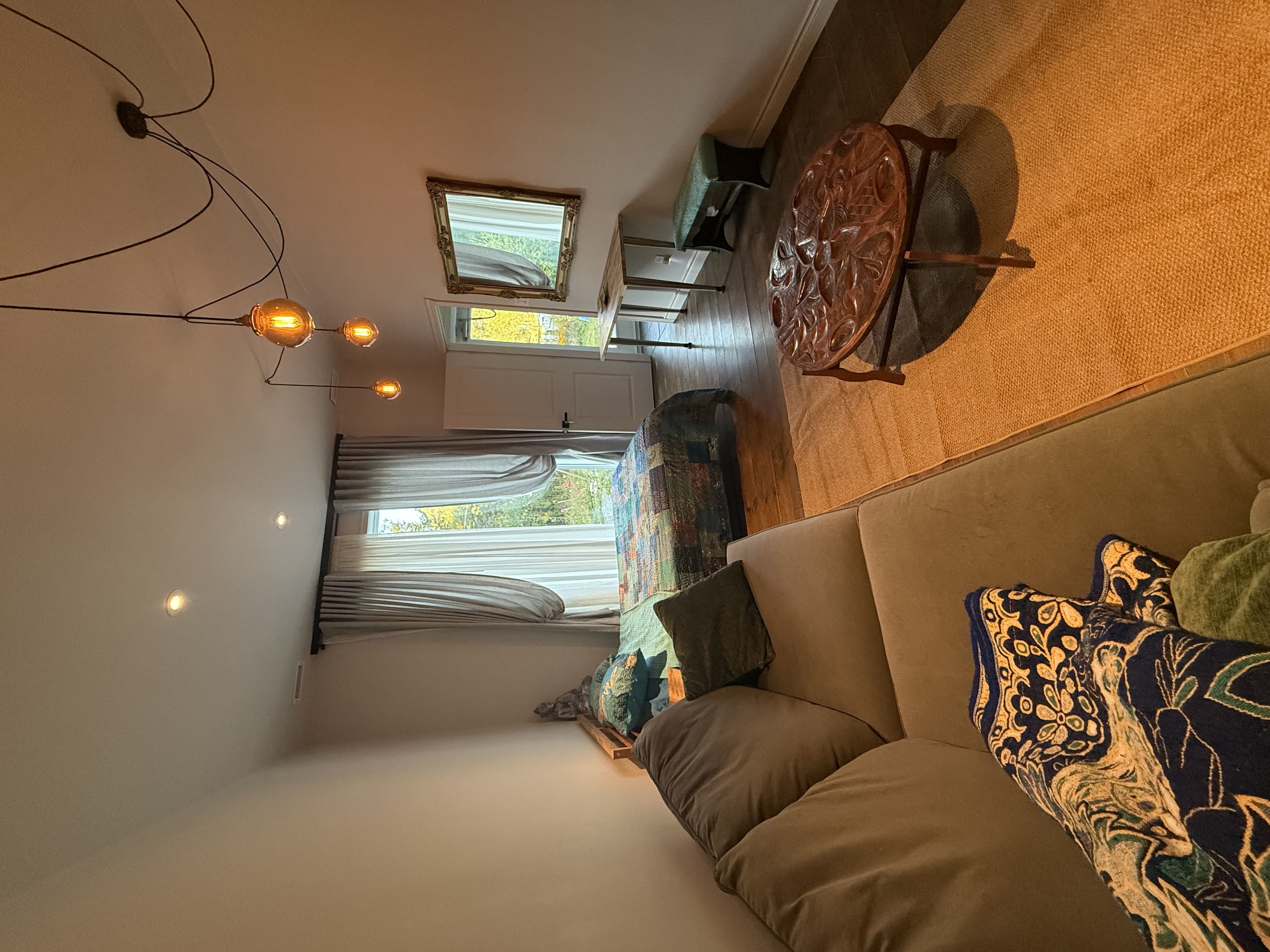 Lit et canapé gris, table basse sculptée en bois, rideaux clairs et éclairage suspendu chaleureux.