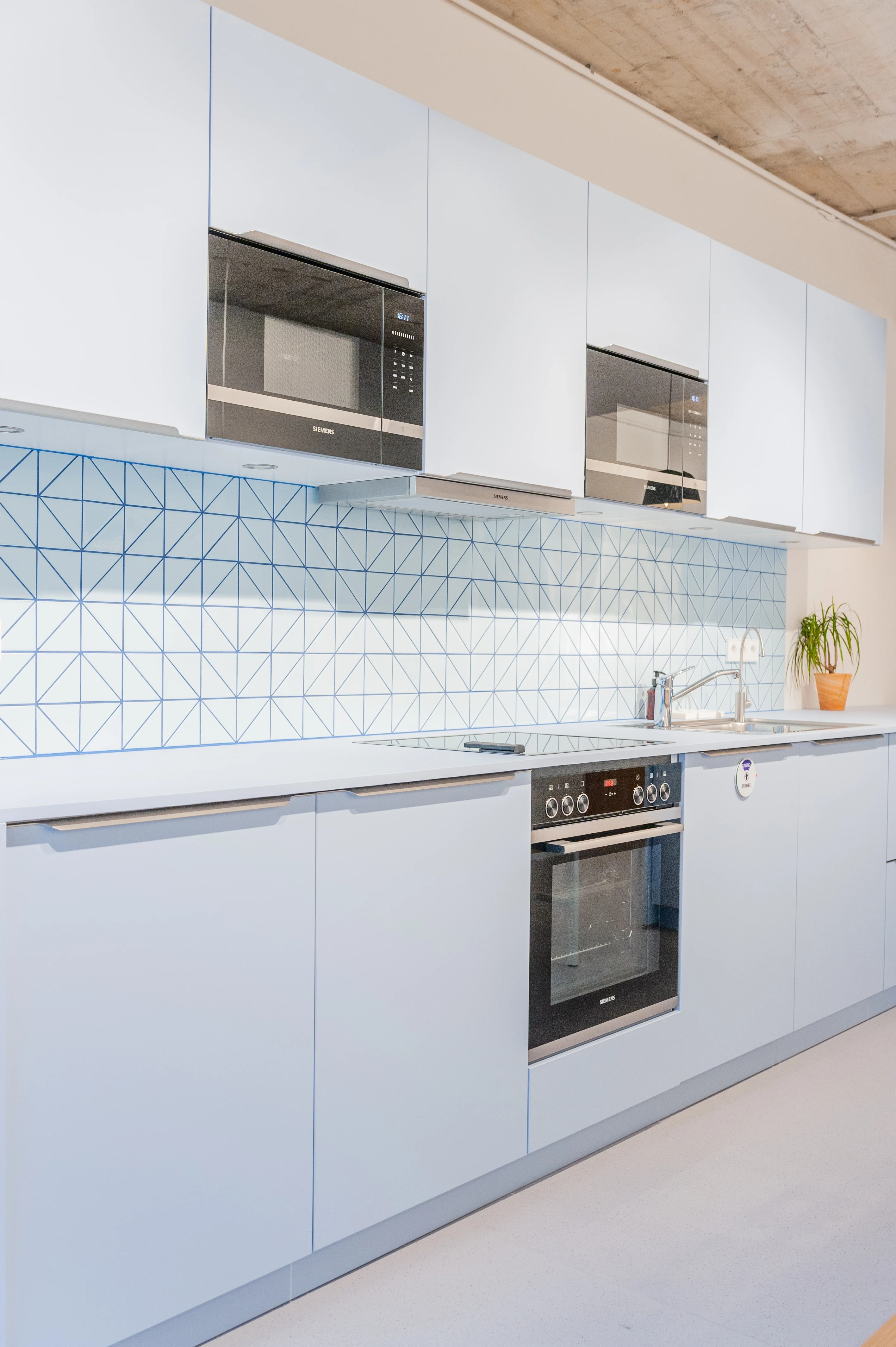 Pale blue kitchen at ZENLOOP with flat cabinet fronts, two microwaves, a geometric blue-line tile splashback, built-in oven and a small potted plant.
