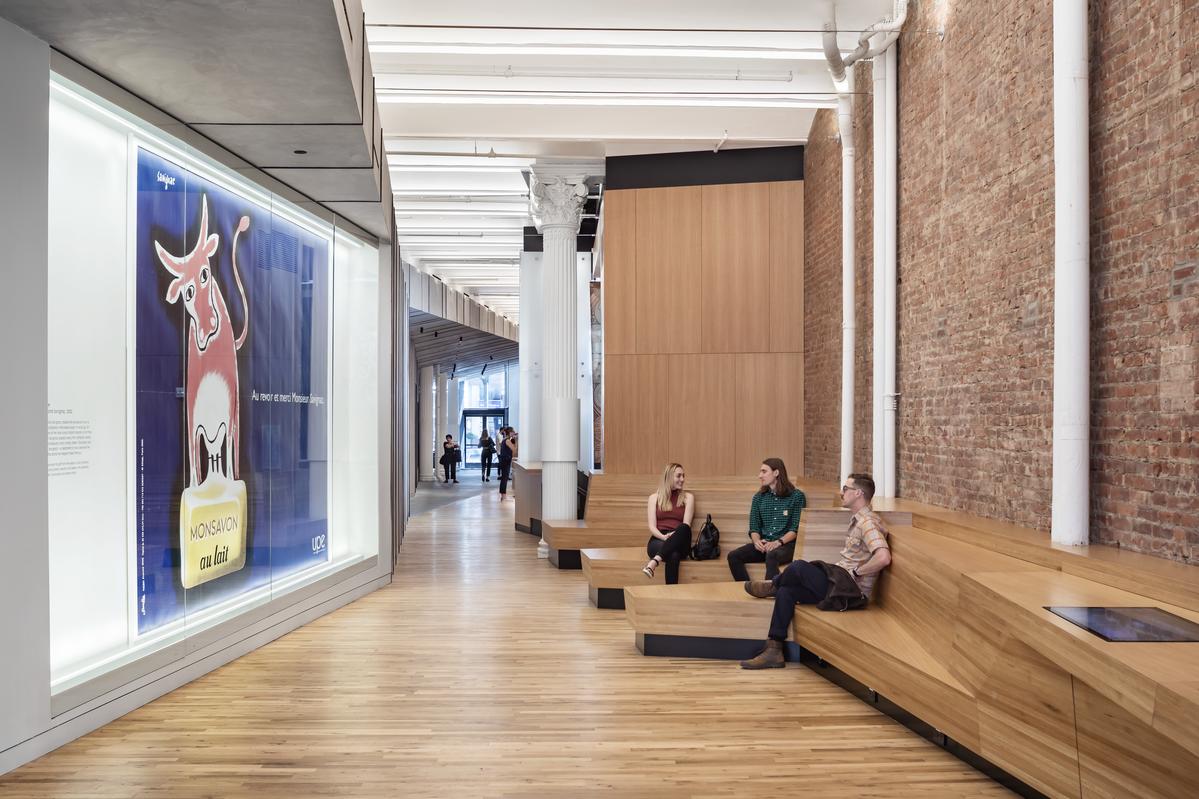 Interior of Poster House in New York City, showing a long wood-floored corridor with a large framed poster on the left wall, built-in wooden seating along an exposed brick wall on the right, and several visitors sitting and walking toward the glass entrance in the distance.