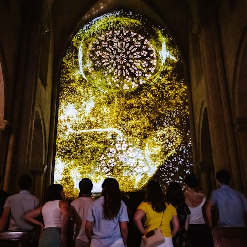 En grupp människor står framför en stor, upplyst skärm med abstrakta gyllene mönster och former inuti en struktur som påminner om en katedral.