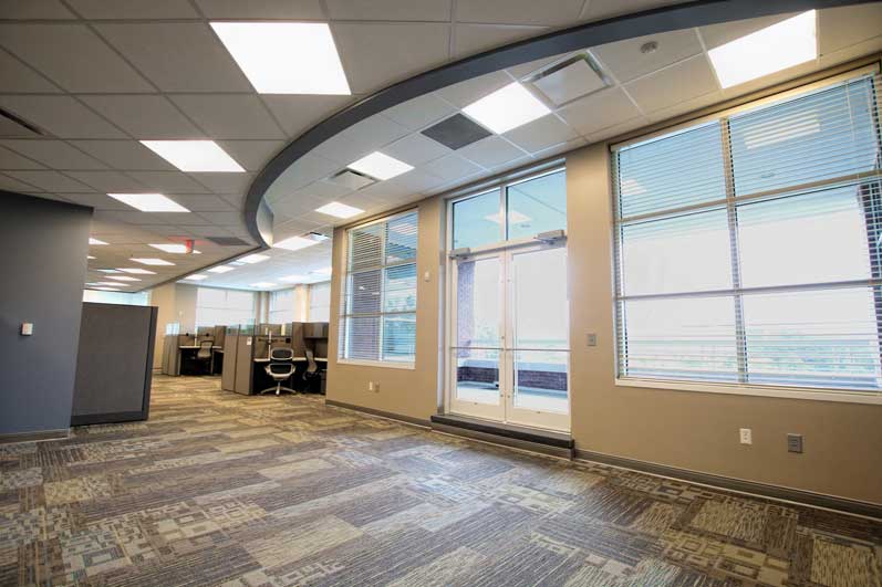 A modern office space with large windows, gray carpet, and curved ceilings, featuring cubicles in the background.