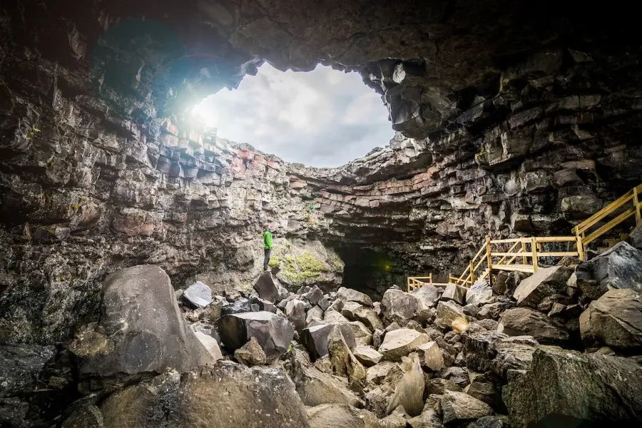 A person in a bright green jacket stands on a boulder inside a massive, layered stone cave, looking up toward a large circular opening in the ceiling that reveals a bright, cloudy sky. Large rocks cover the cave floor, and a wooden staircase descends into the cavern on the right.